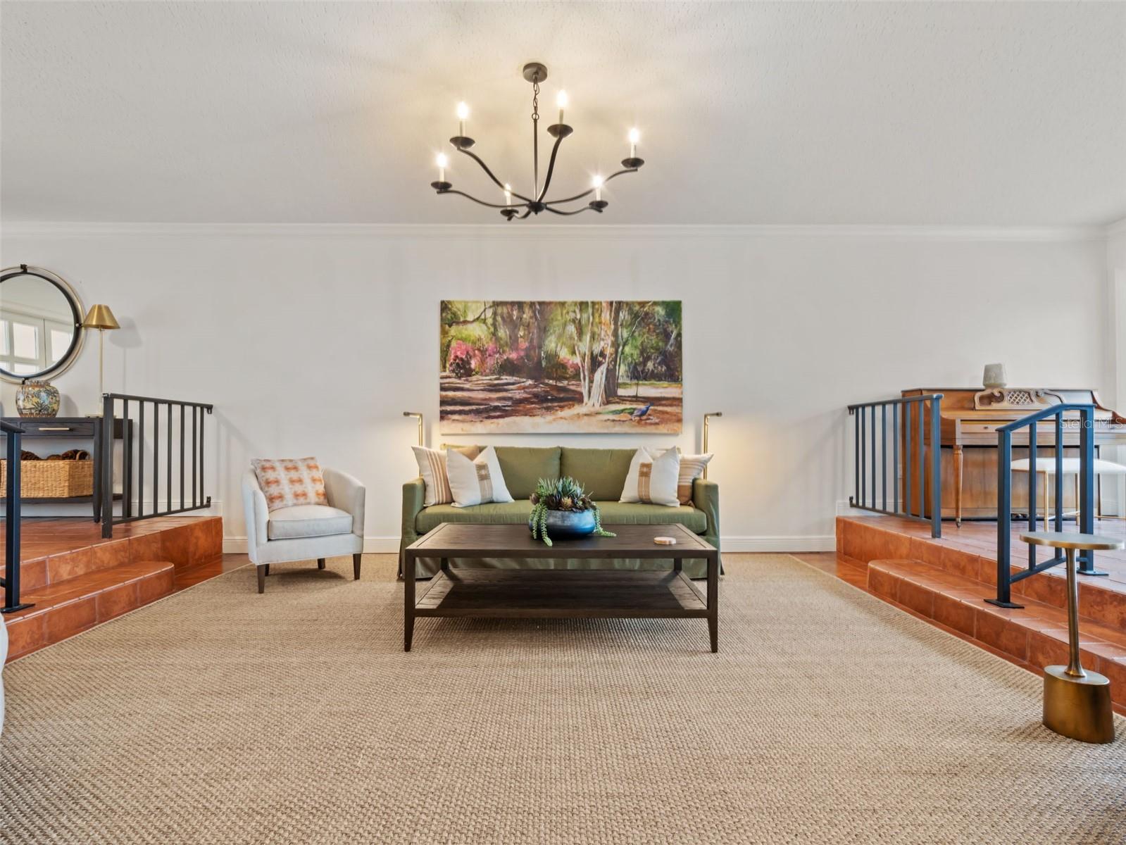 Staged living room with a green sofa, woven rug, chandelier, and terracotta-tiled raised areas.