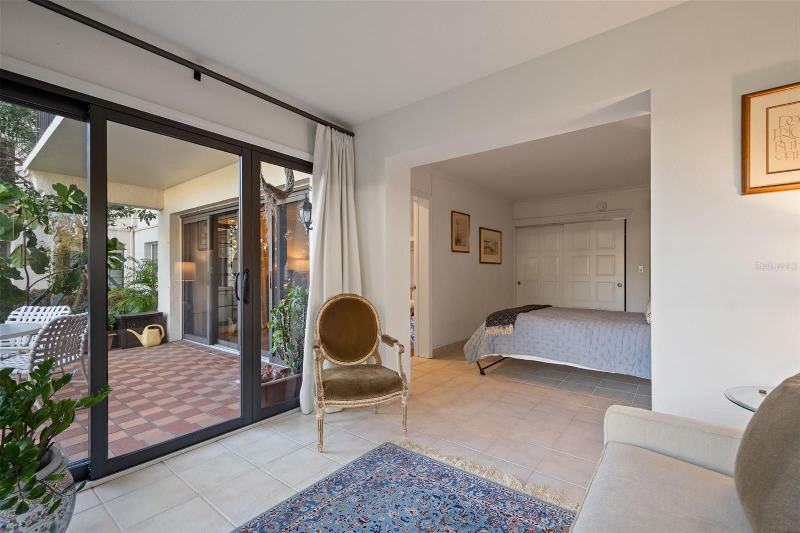 Bedroom with sliding glass doors opening onto a tiled patio with plants and outdoor seating.