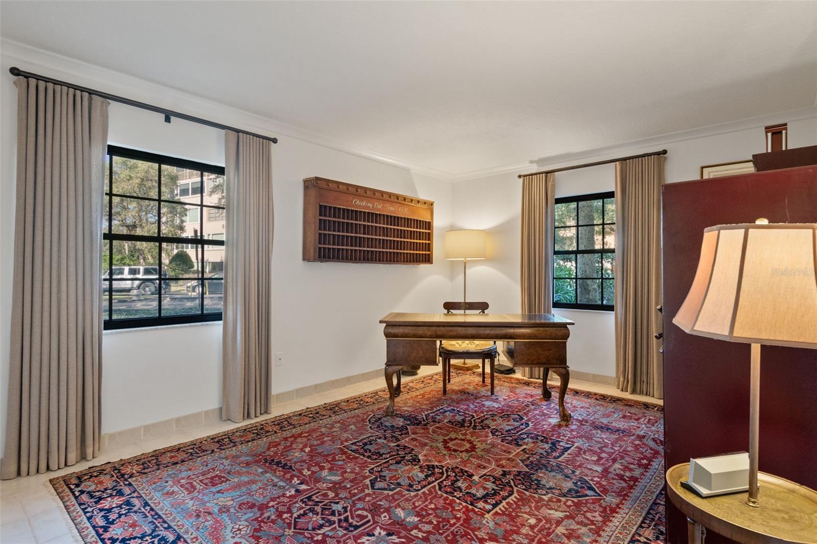 Furnished office with an antique wooden desk, patterned area rug, and curtained windows.