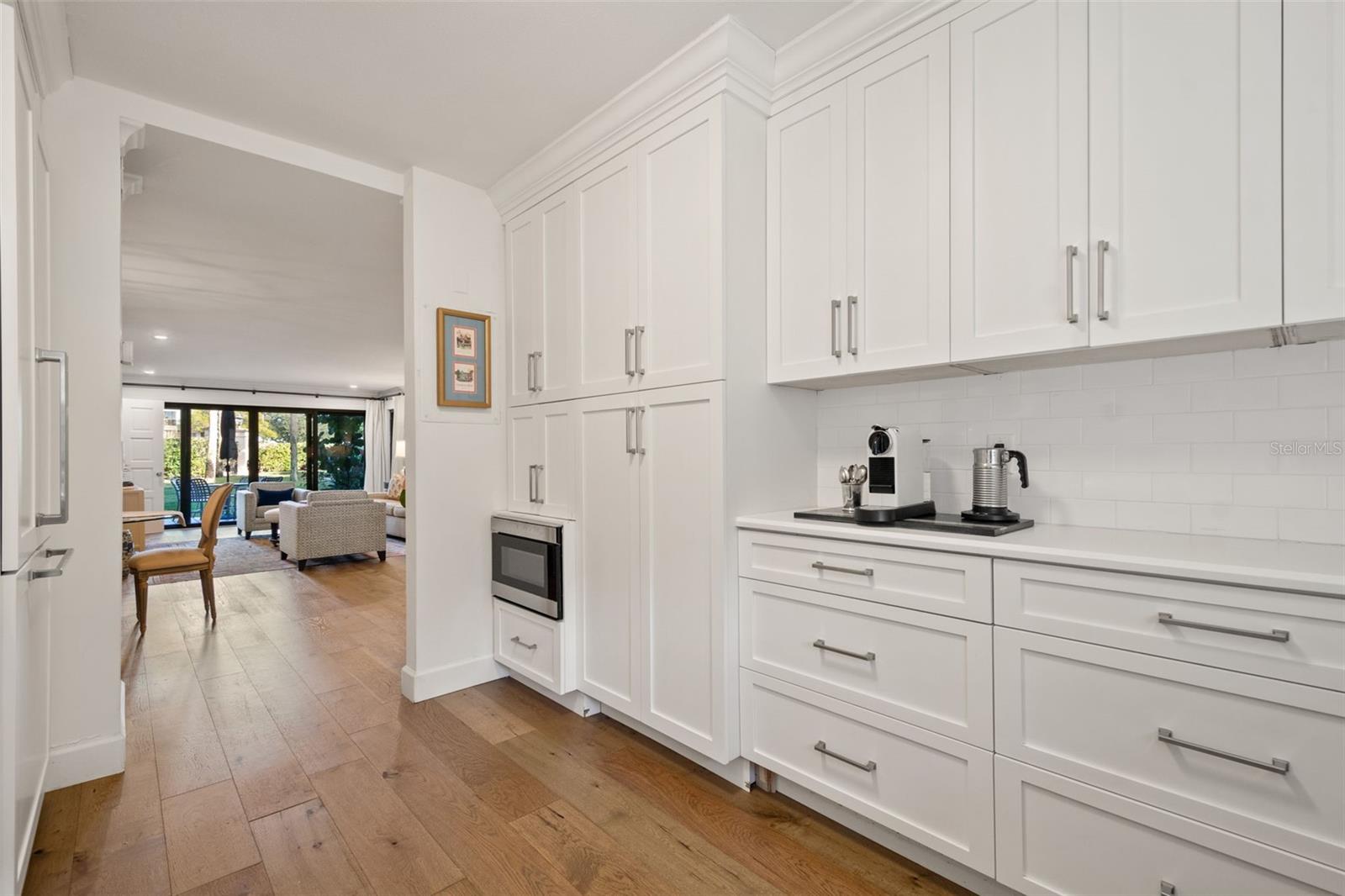 White-cabinet kitchen with wood floors, opening to a furnished living room and glass doors.