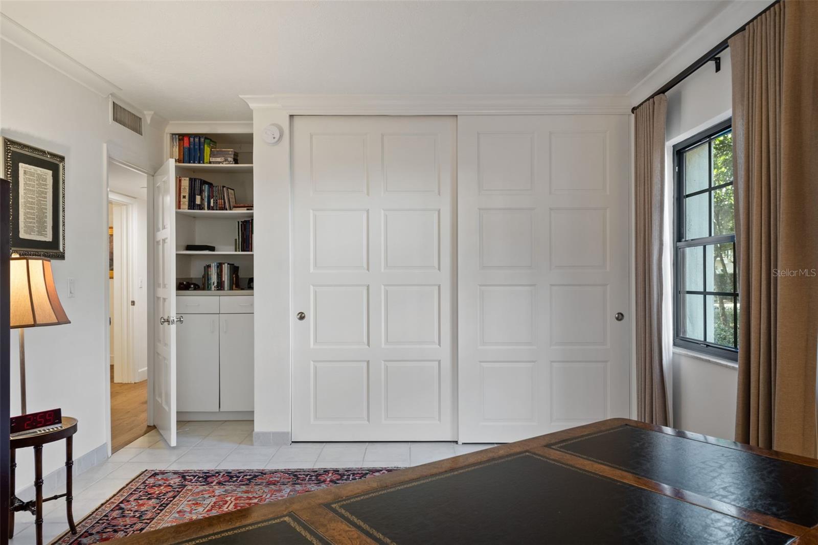 Bedroom with paneled closet doors, built-in bookshelves, and a curtained window beside a desk.