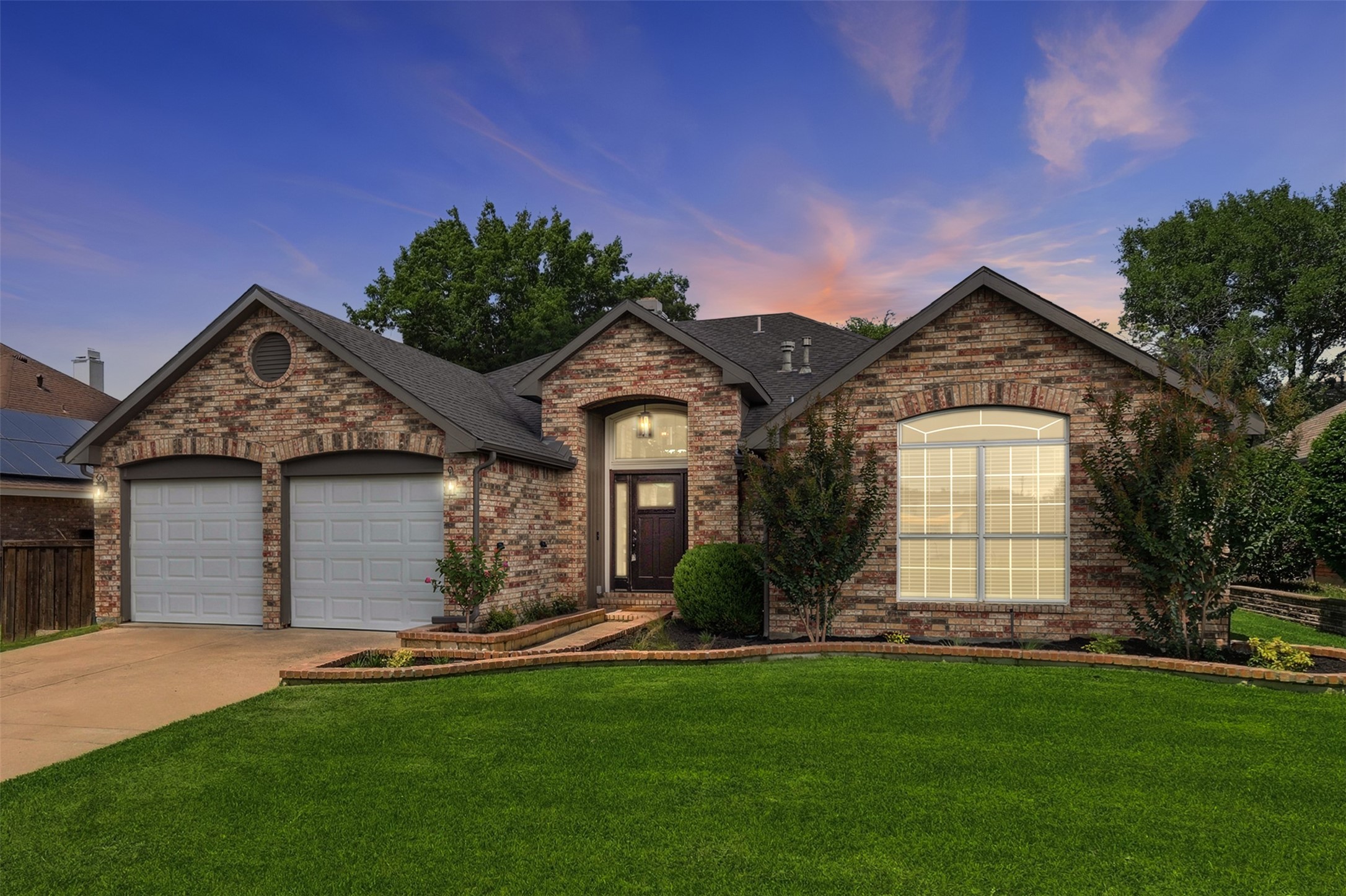 Brick house with two garage doors and a broad lawn beneath a colorful dusk sky.