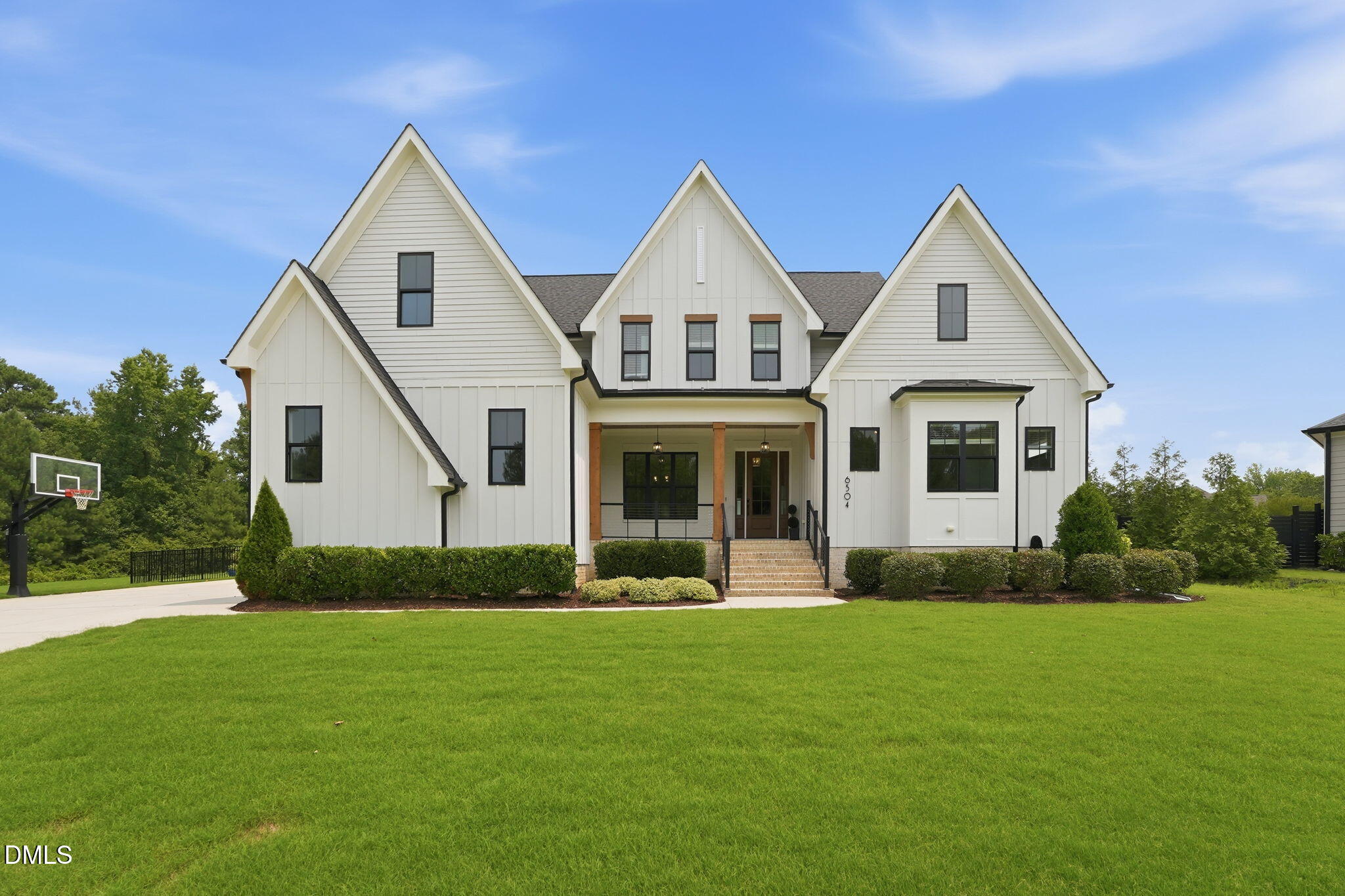 White multi-gable house with a covered porch, brick steps, trimmed hedges, and a basketball hoop by the driveway.