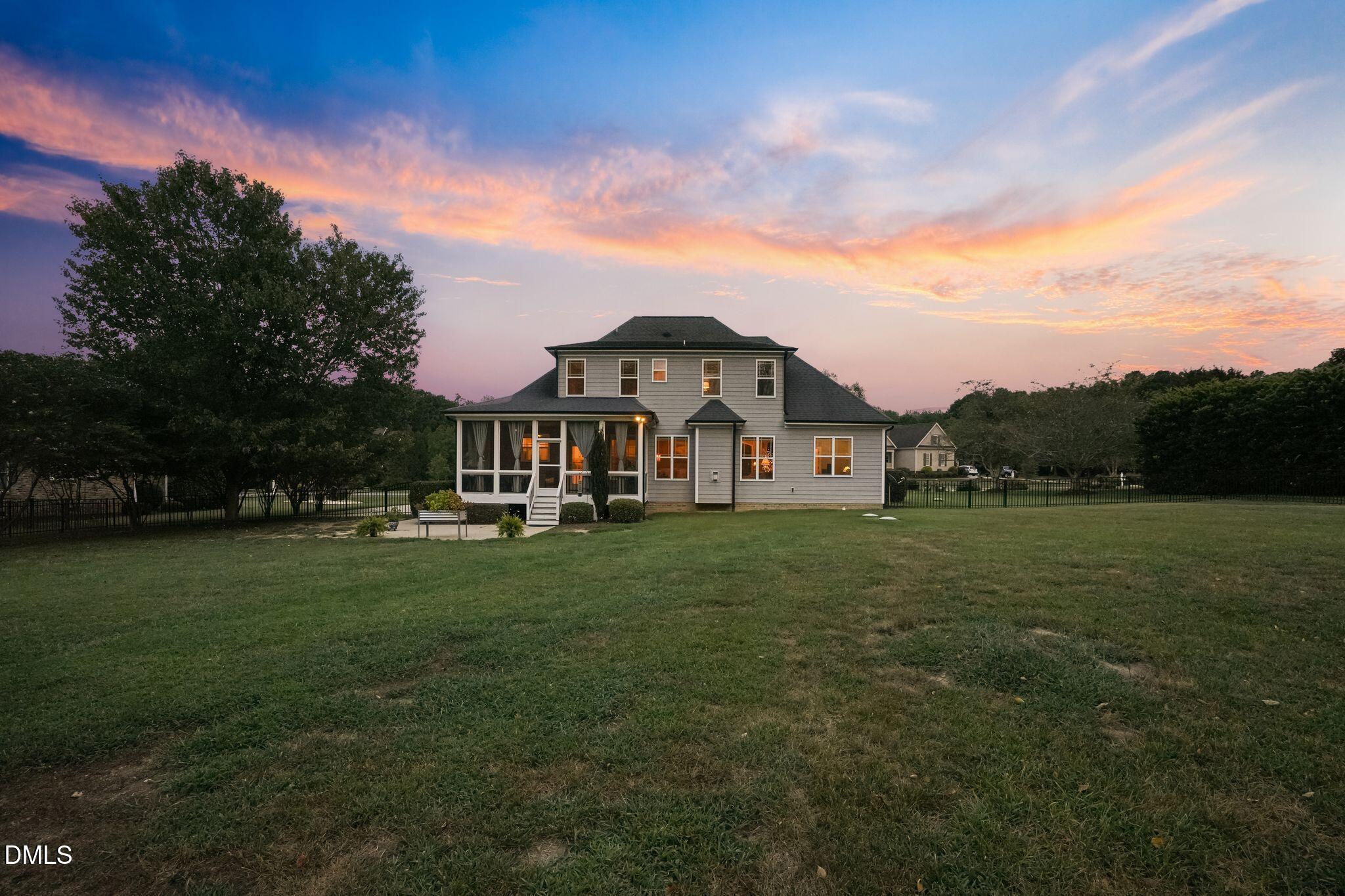 Two-story gray house with a screened porch and broad fenced lawn at sunset.