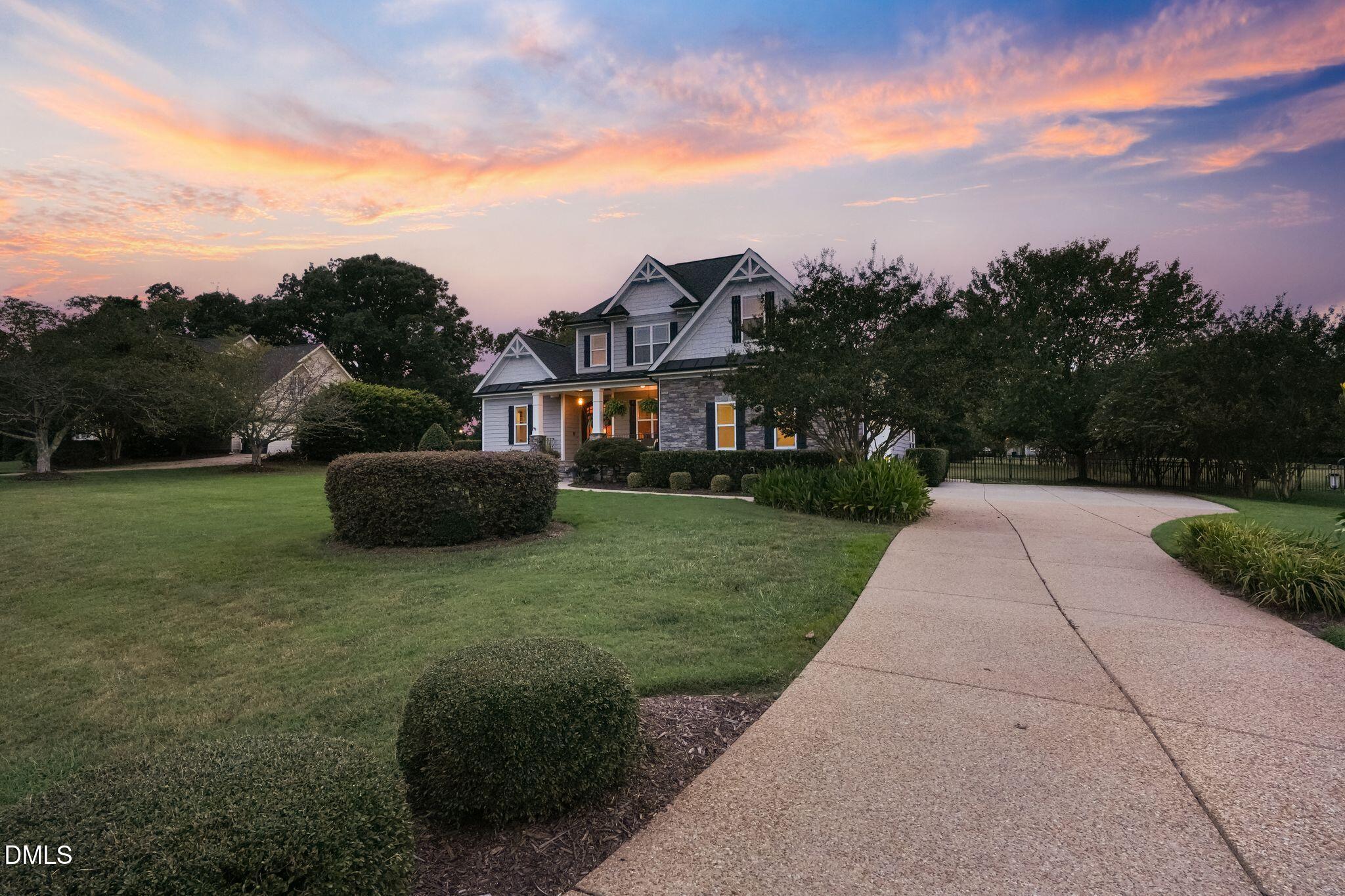 Two-story gray house with stone facade and curved driveway beneath a pink and orange sunset sky.