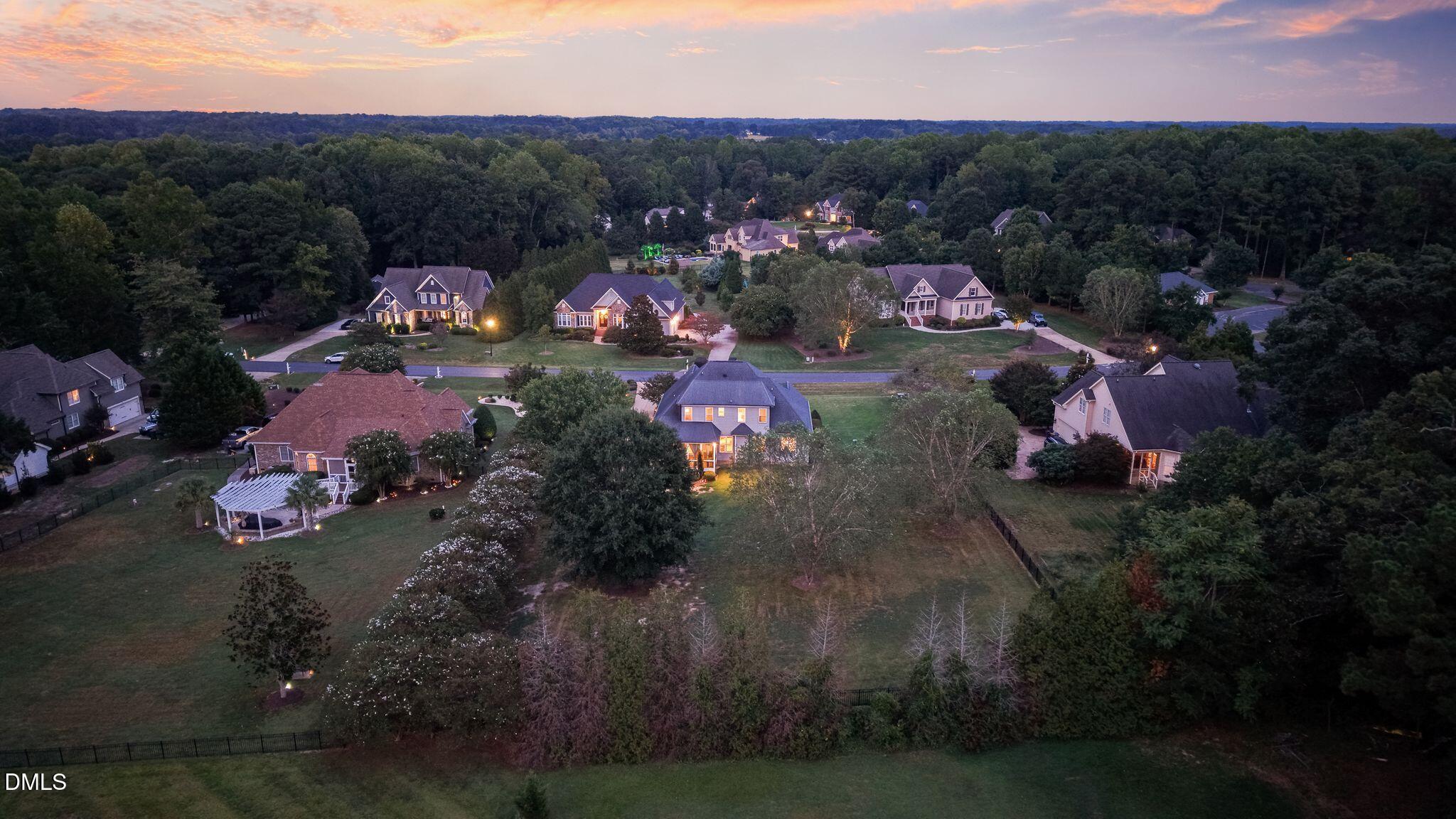 Aerial view of homes among trees at dusk, with glowing windows and a pink-orange sky.