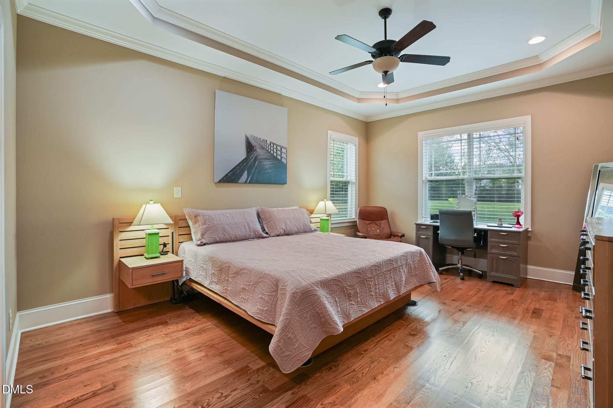 Bedroom with a large bed, hardwood floors, tray ceiling and a desk workspace by the windows.