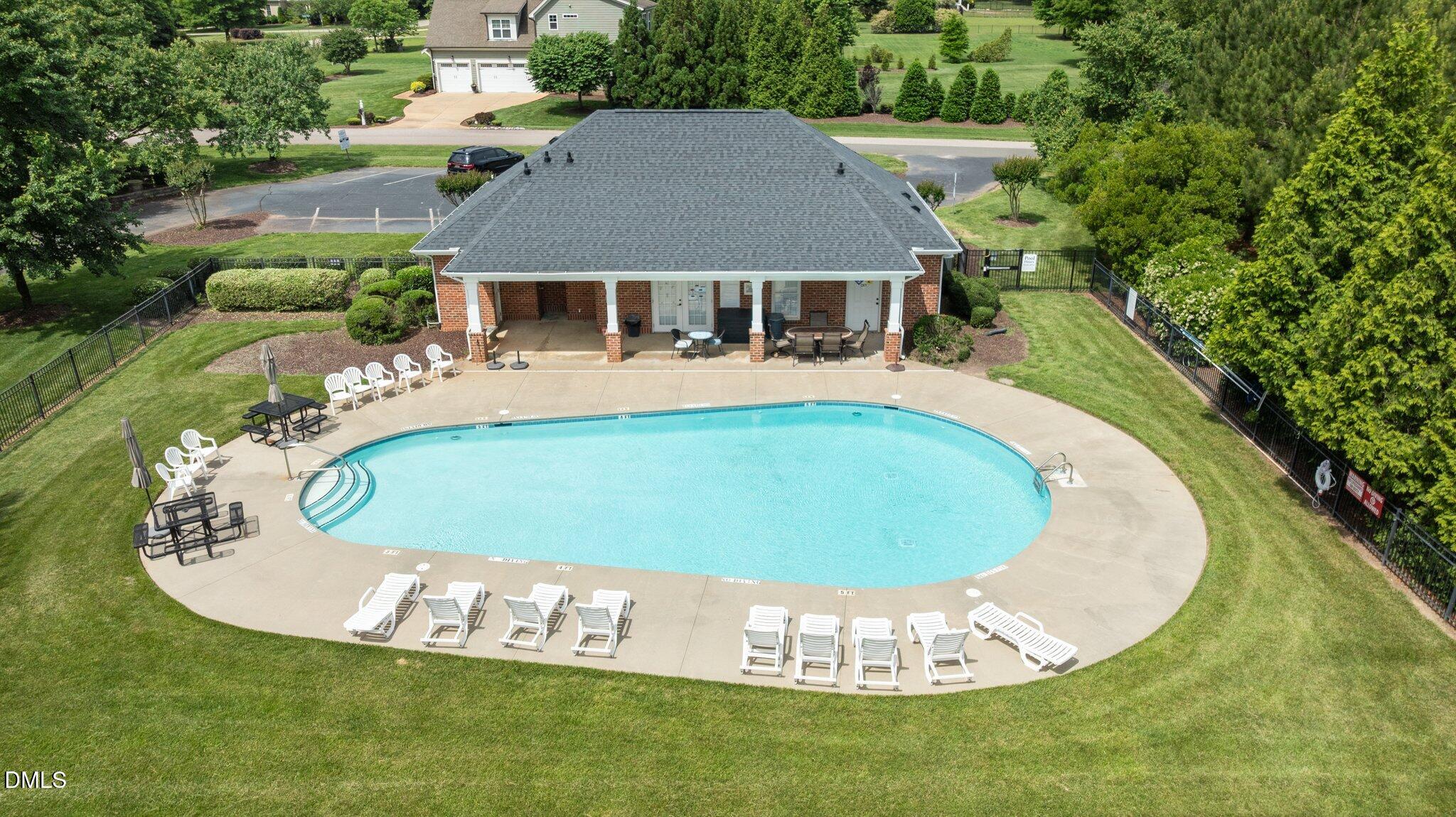 Aerial view of a swimming pool and brick pavilion, surrounded by lounge chairs and a fenced lawn.