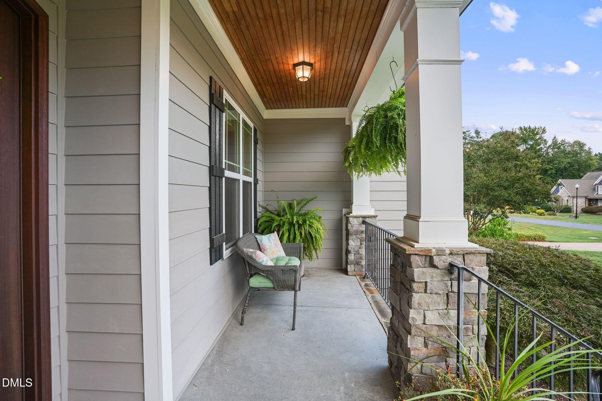 Covered front porch with a wood ceiling, stone column bases, hanging fern, and cushioned wicker chair.