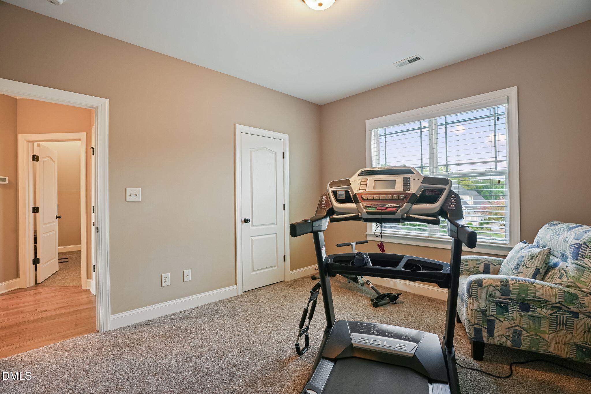 Home workout room with a treadmill, large window, and patterned armchair.