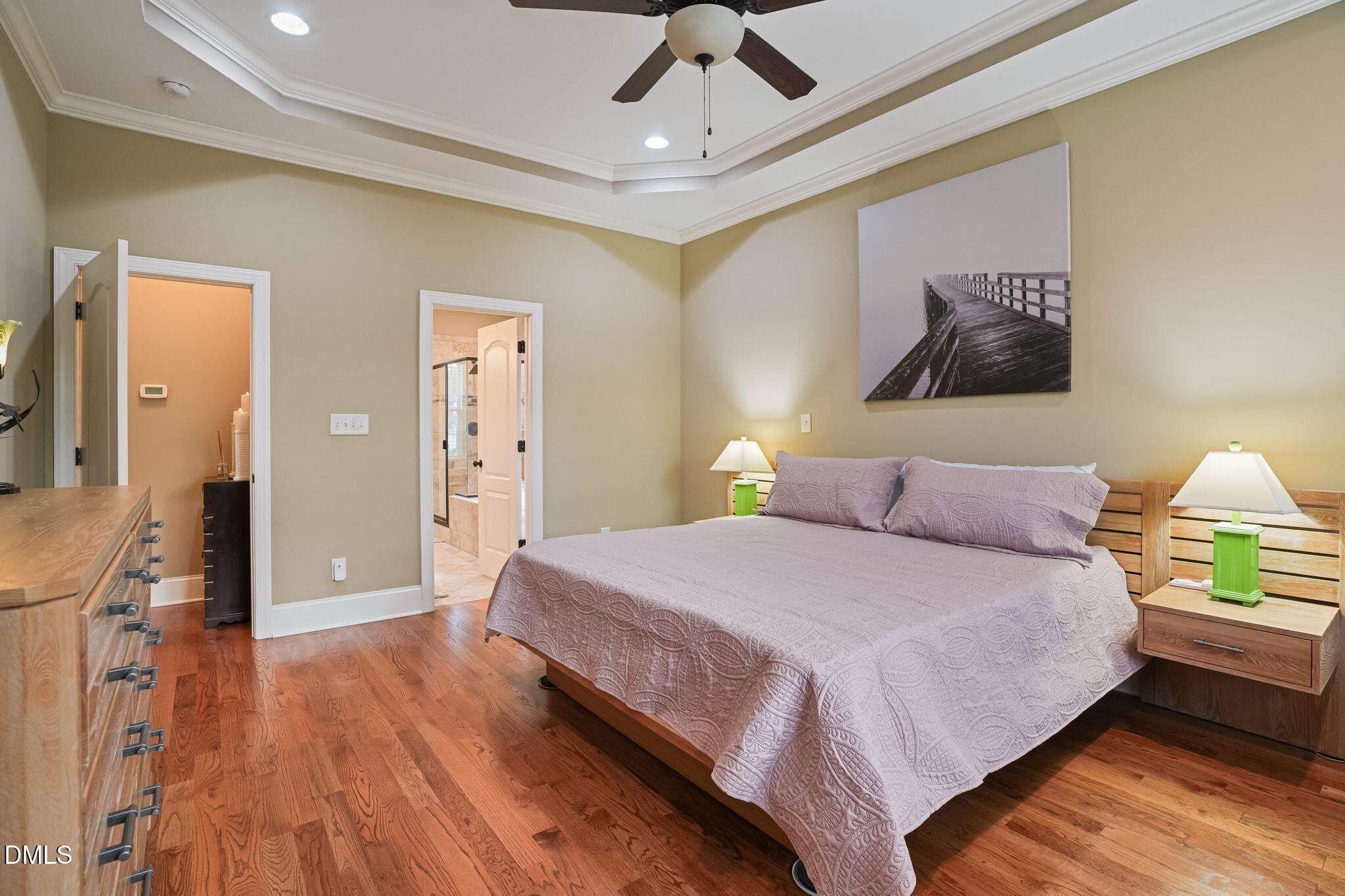 Furnished bedroom with a large lavender-covered bed, hardwood floors, and a tray ceiling with ceiling fan.