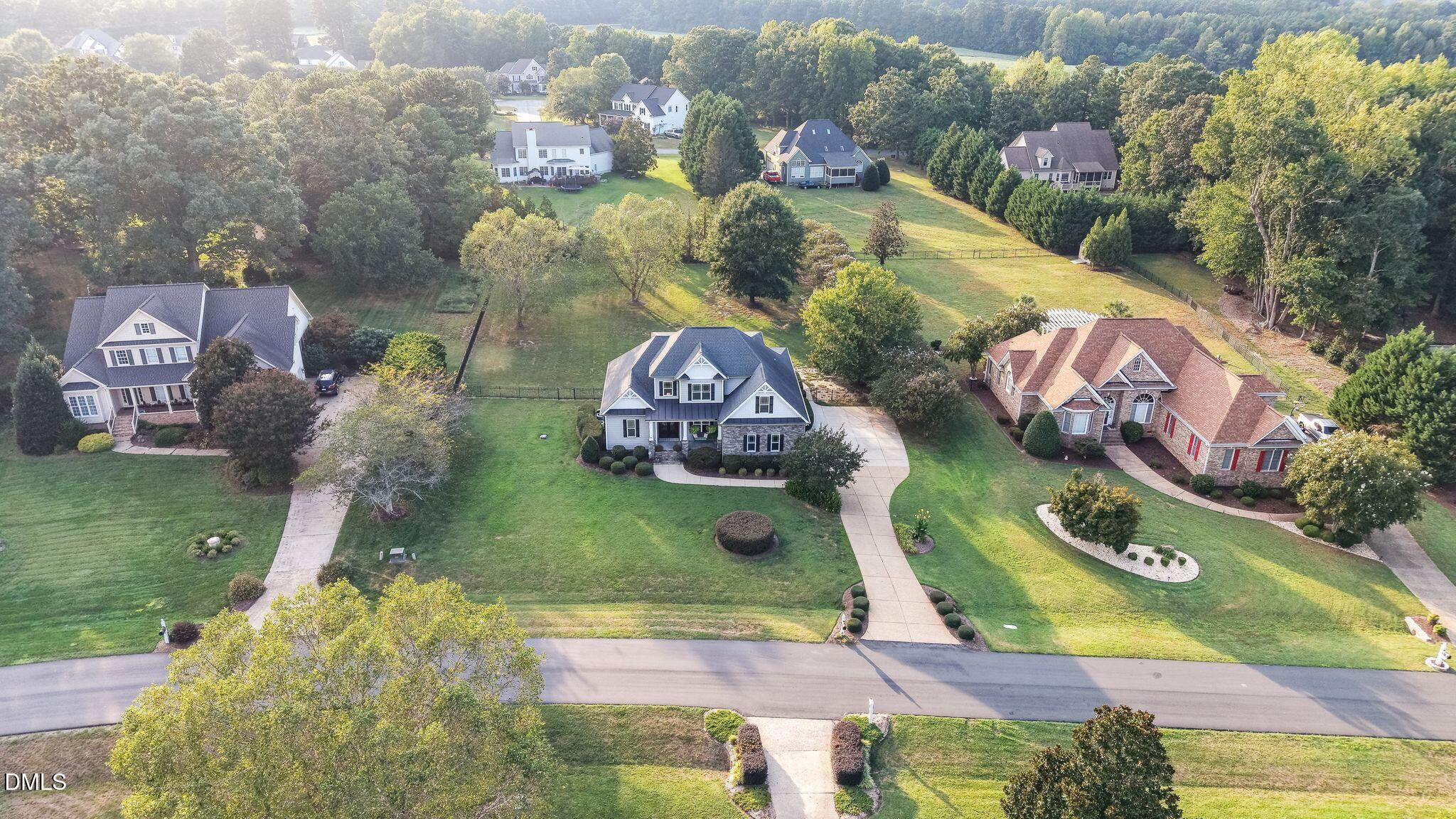Aerial view of a gray-and-white home with a curved driveway, neighboring houses, and wooded backdrop.