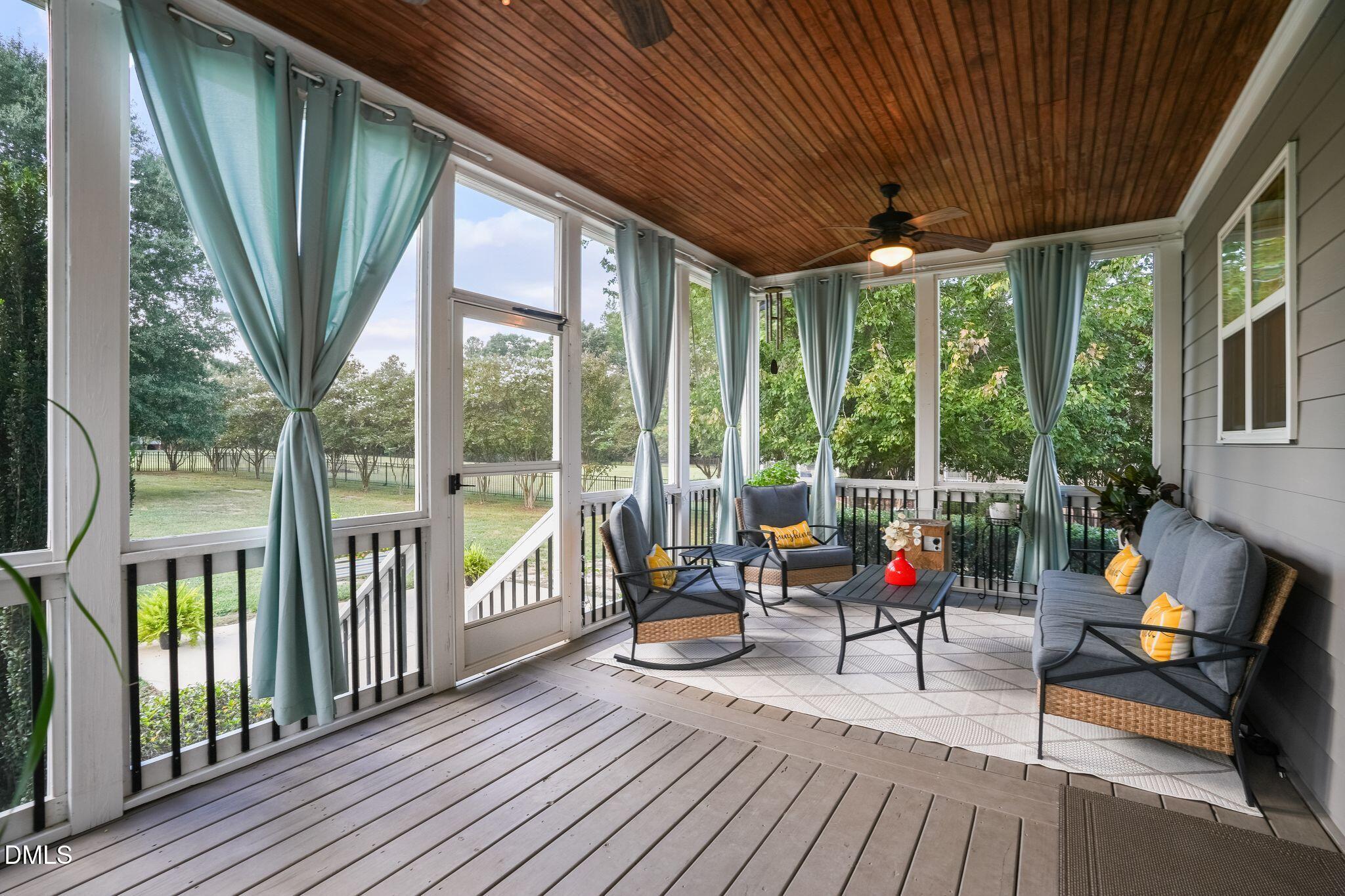 Screened porch with a wood ceiling, pale blue curtains, and cushioned seating overlooking a leafy yard.