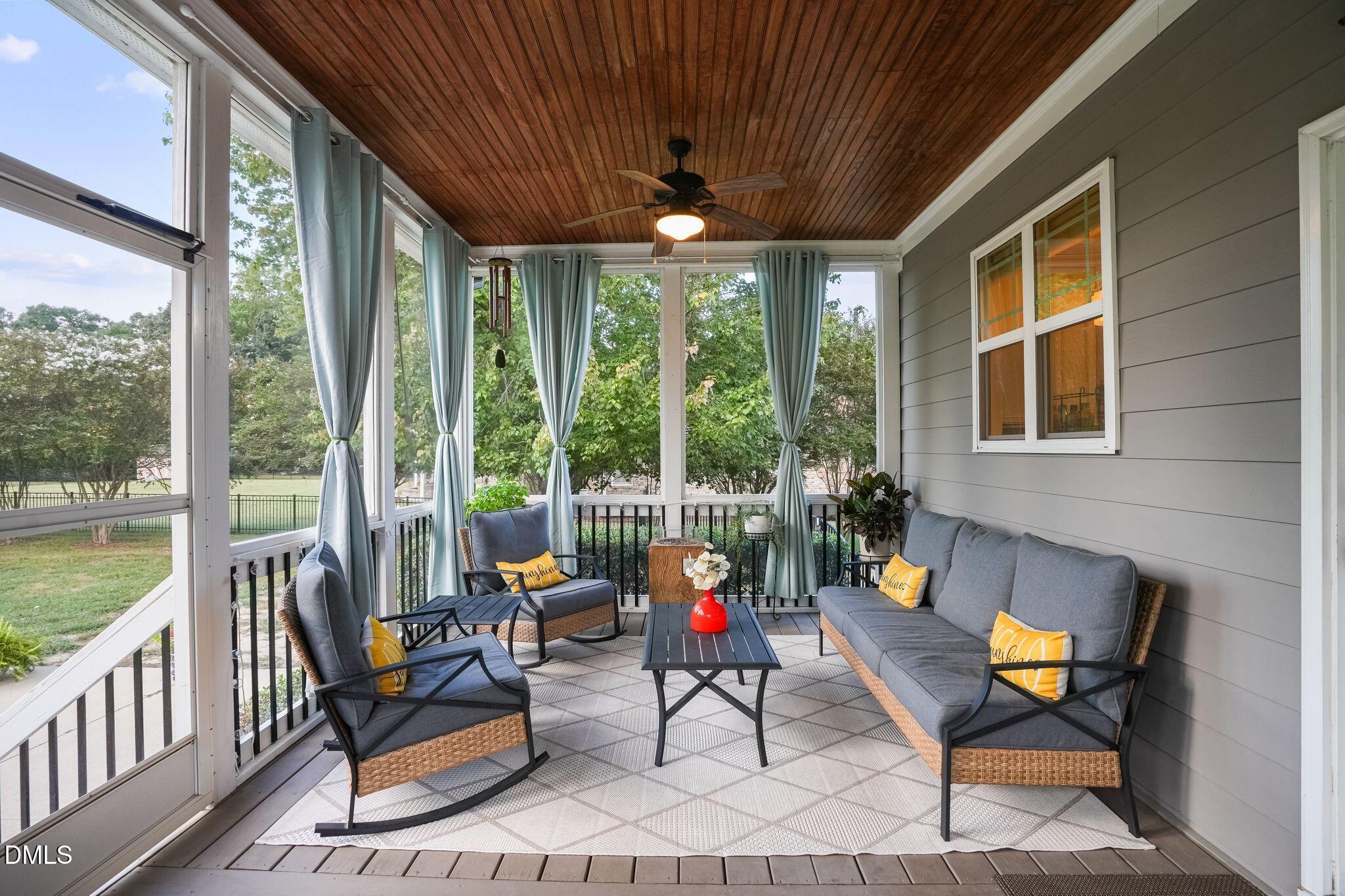 Screened porch with gray seating, blue curtains, and a wood-plank ceiling overlooking a leafy yard.
