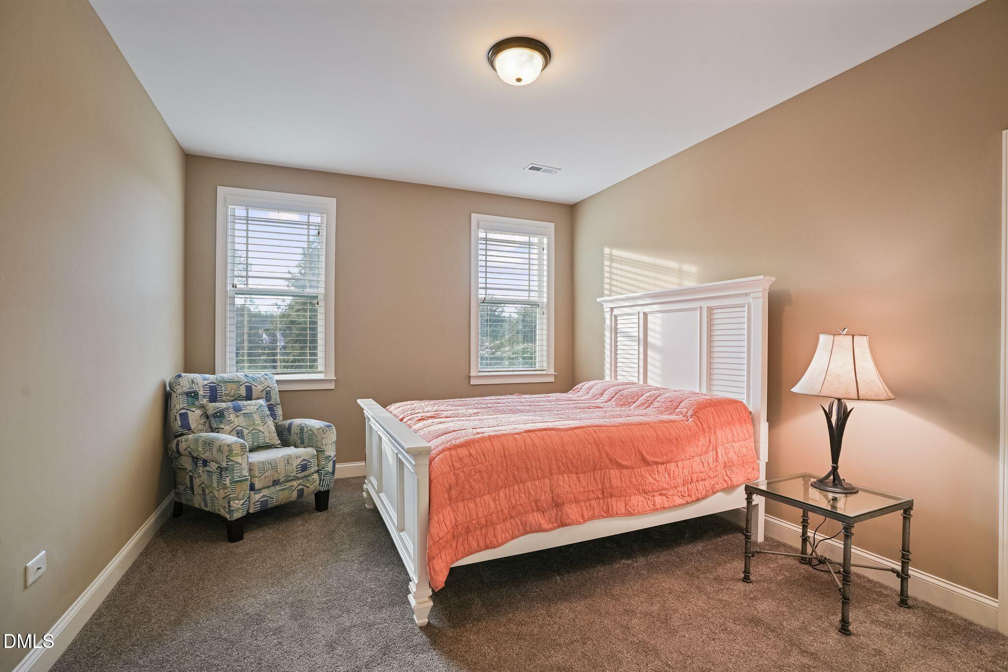 Bedroom with a white paneled bed, coral quilt, two blind-covered windows, and a patterned armchair.