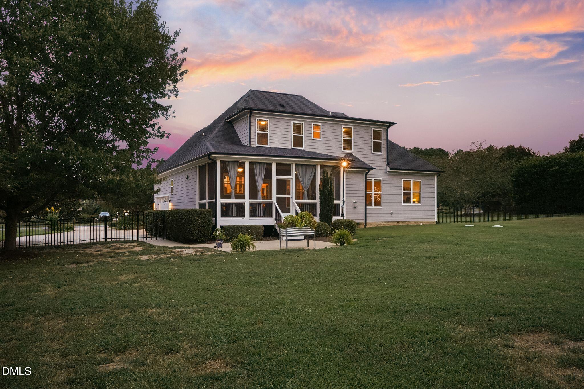 Two-story gray home with a screened porch and fenced lawn beneath a colorful dusk sky.