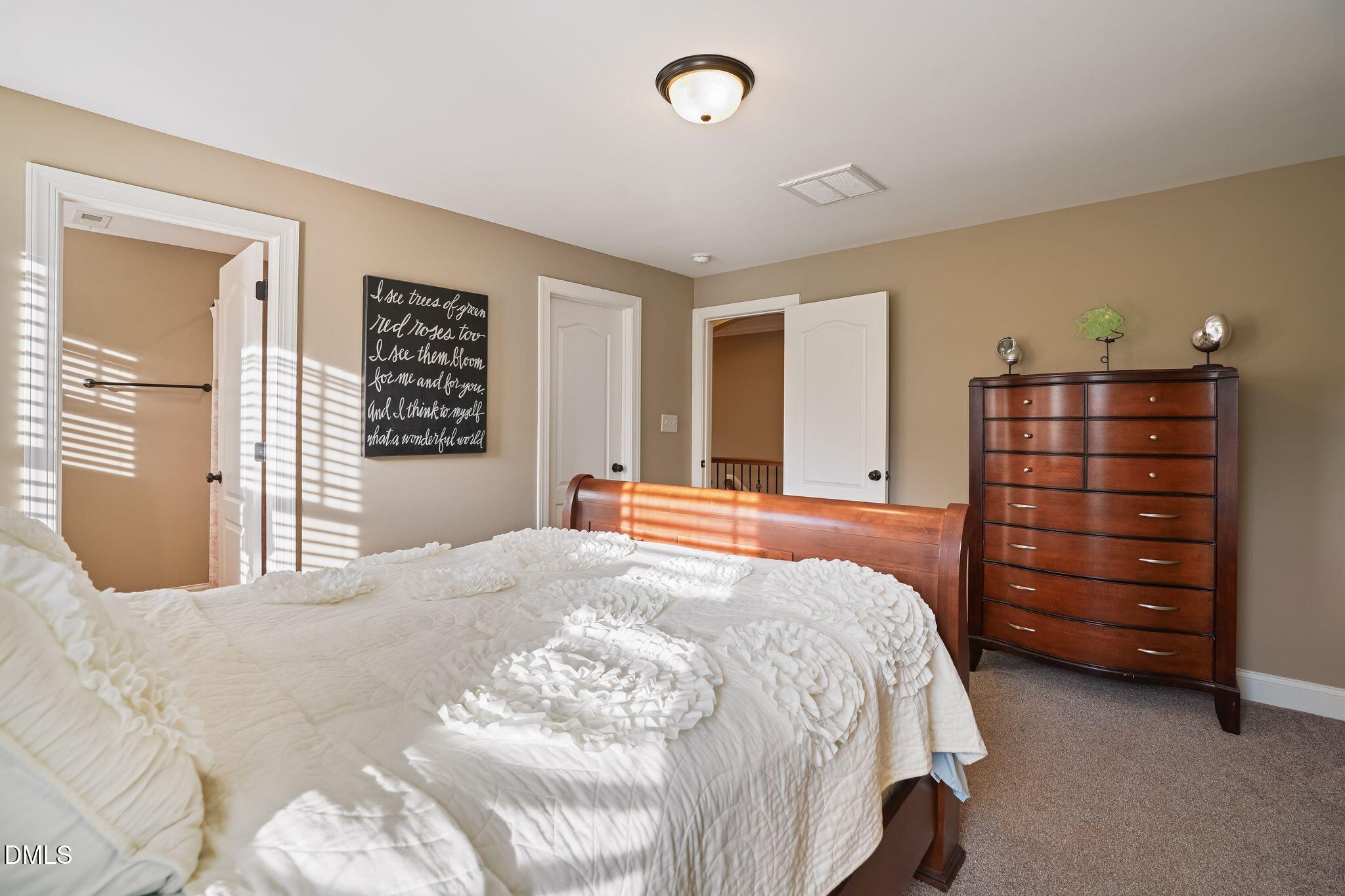 Bedroom with white ruffled bedding, a dark wood dresser, and sunlight striped across the room.