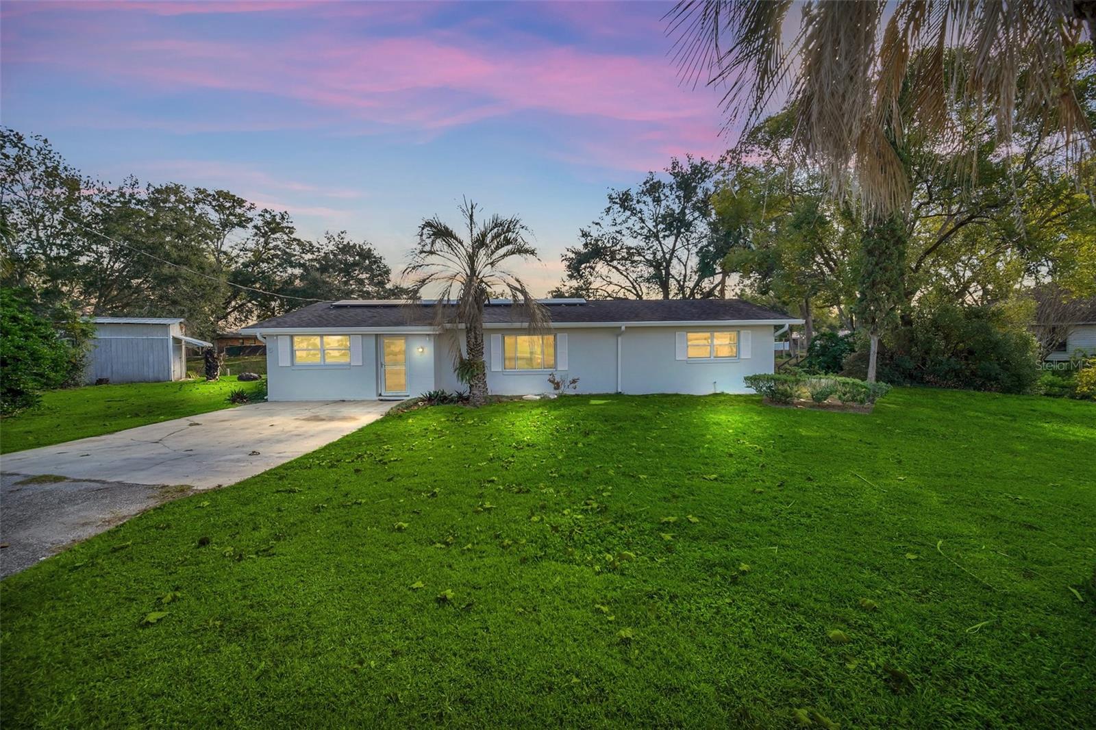 Single-story home with a broad lawn, driveway, palm trees, and a pink-and-blue dusk sky.