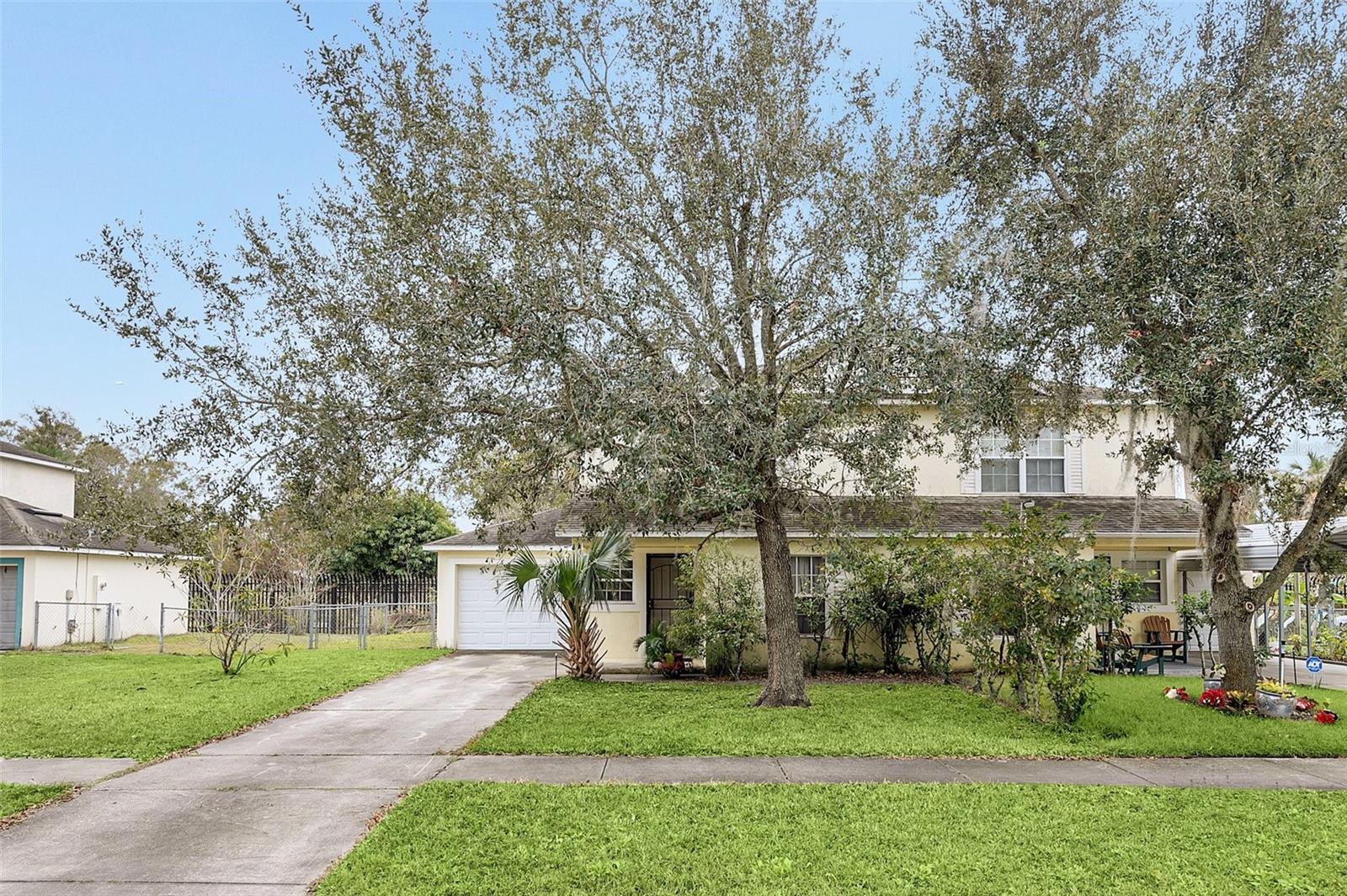 Two-story cream house partly shaded by a mature oak, with an attached garage and concrete driveway.