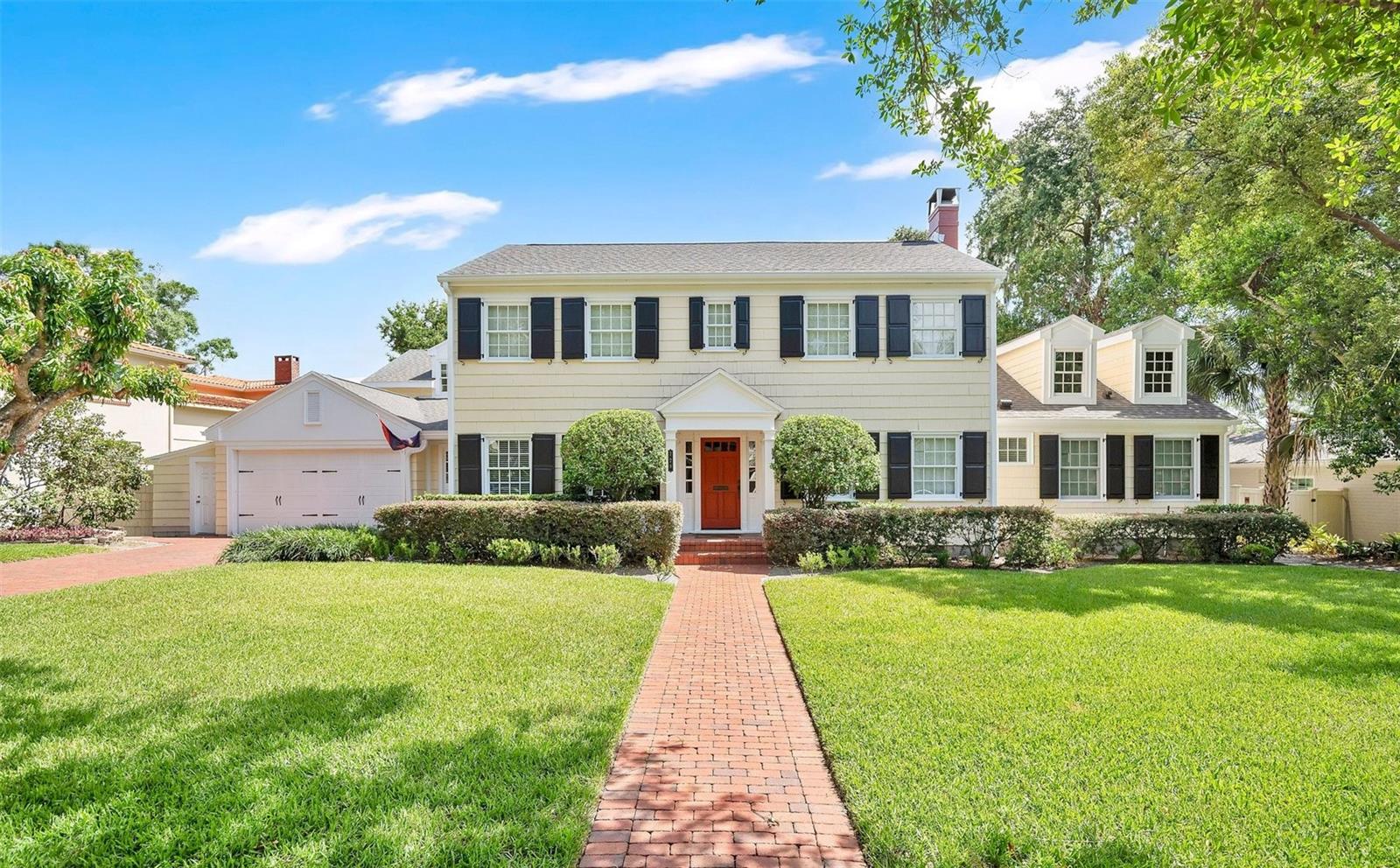 Two-story cream house with black shutters, a red front door, and a brick walkway across the lawn.
