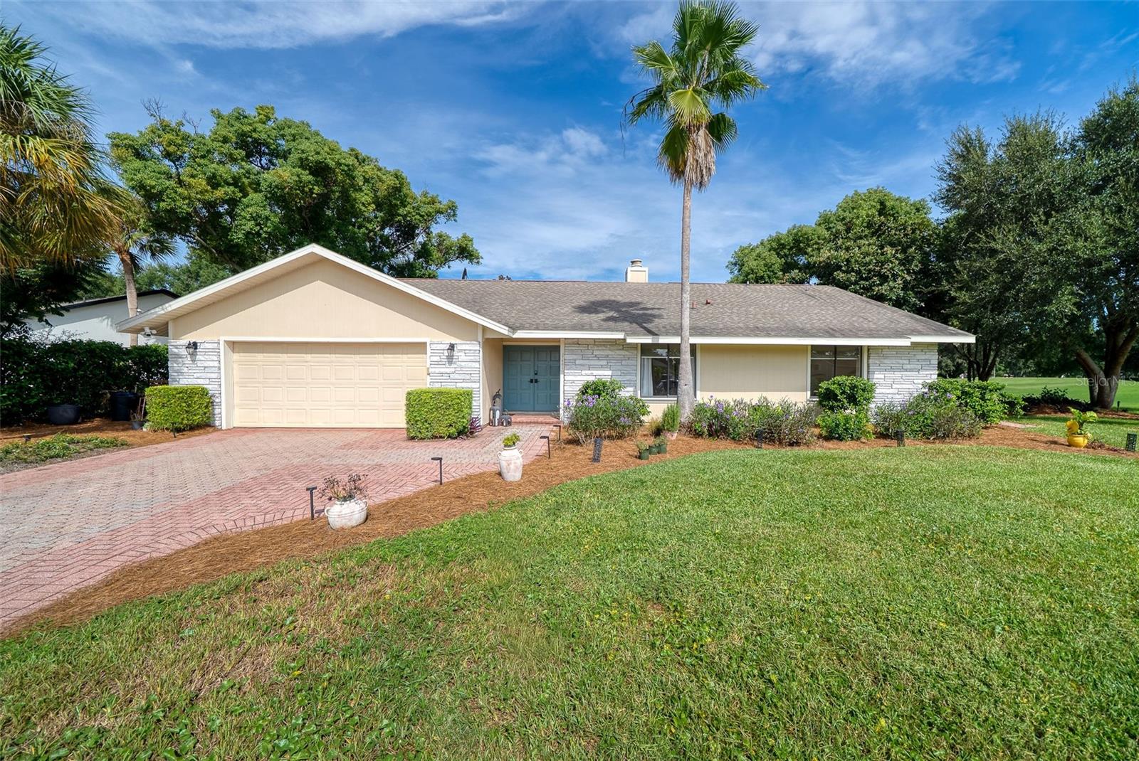 Single-story home with a stone facade, two-car garage, brick driveway, and palm tree in front.