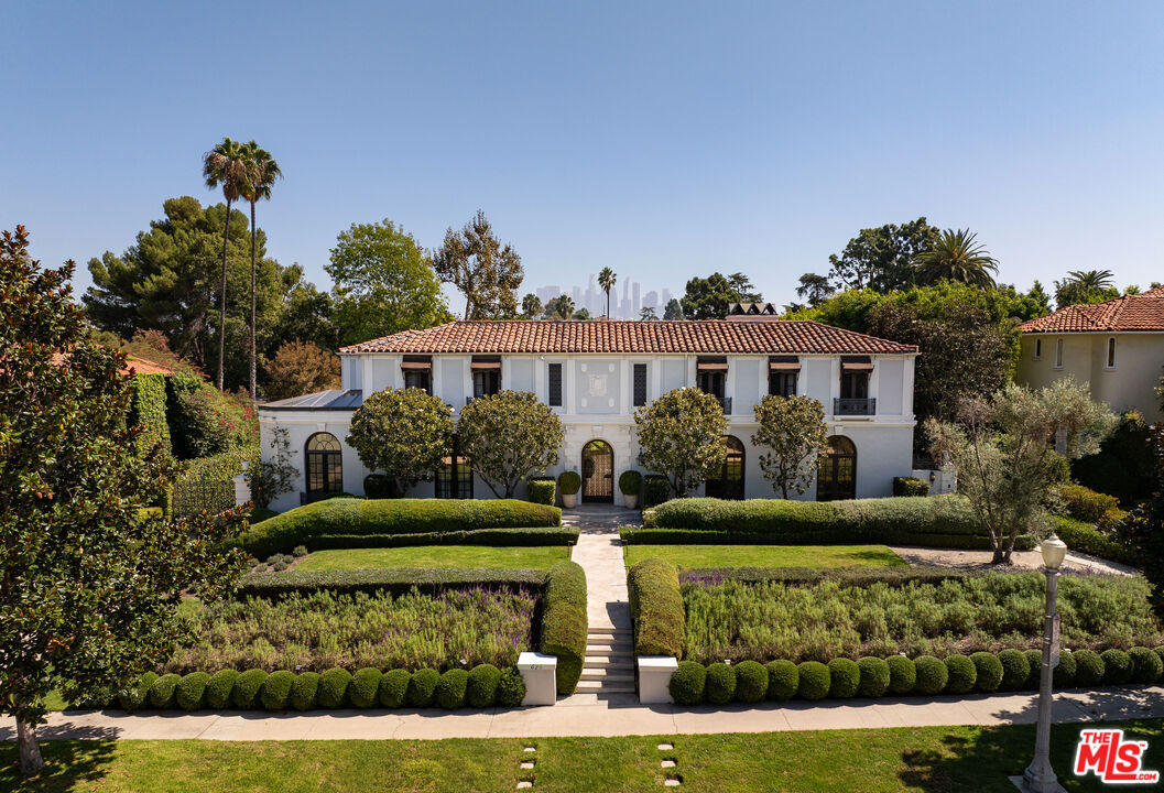 White two-story home with a red tile roof, formal clipped hedges, and a central garden walkway.