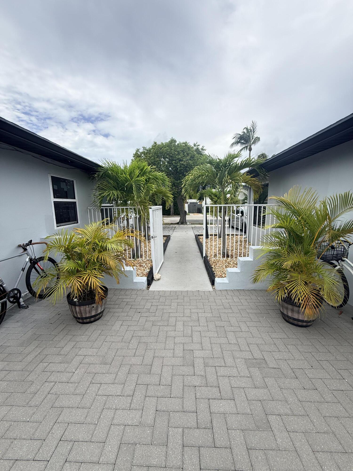 Paved entrance walkway between buildings, bordered by white railings and potted palms.
