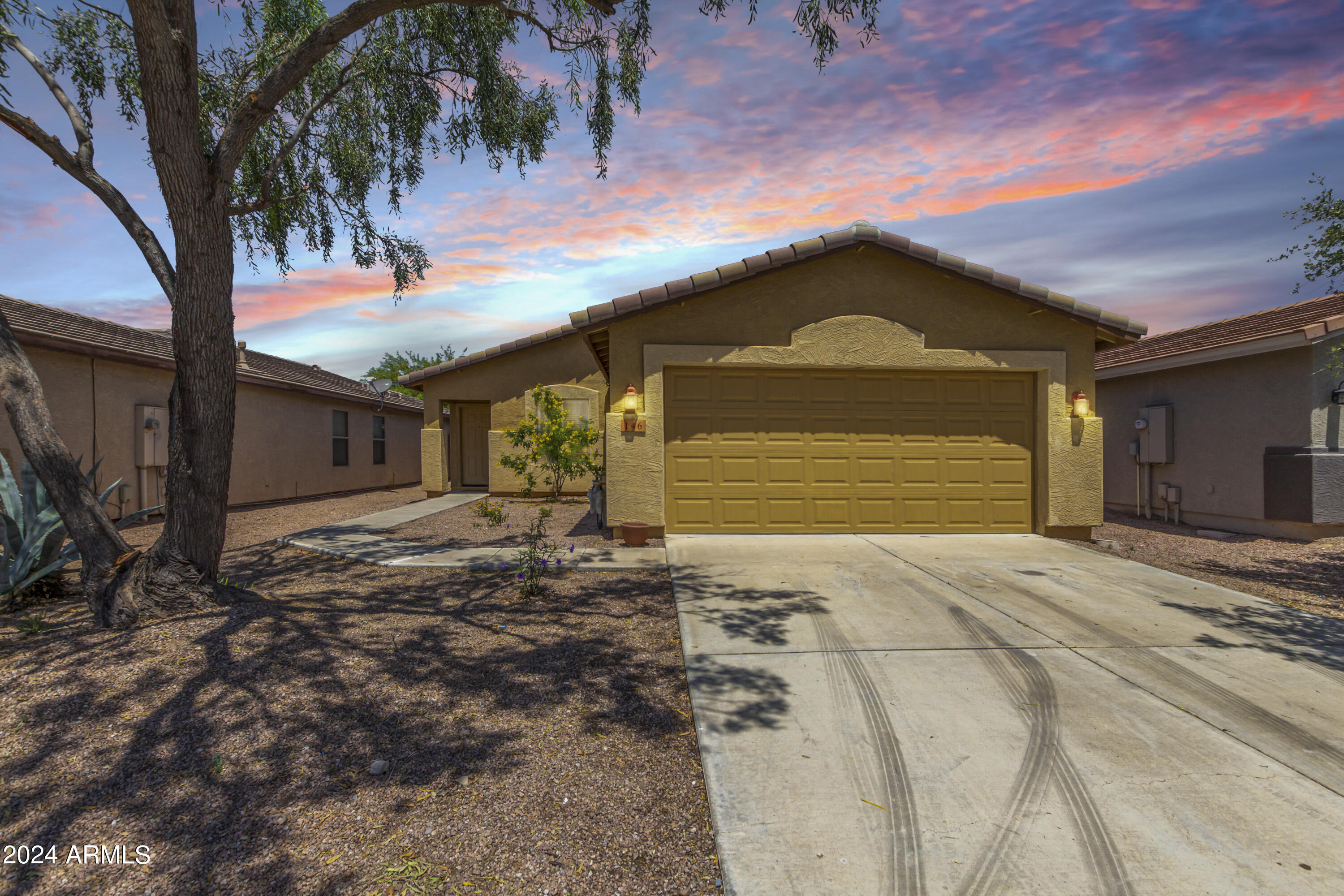 Stucco home with a tiled roof, two-car garage, gravel landscaping, and a large shade tree at sunset.