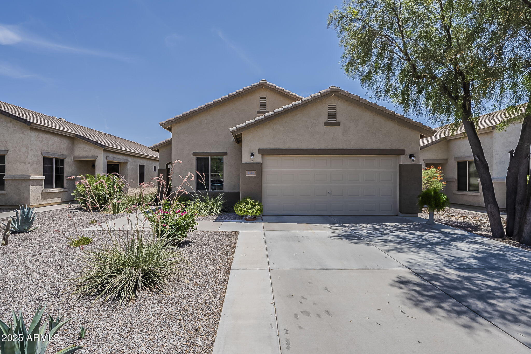 Stucco home with a two-car garage, desert landscaping, and a large shade tree.