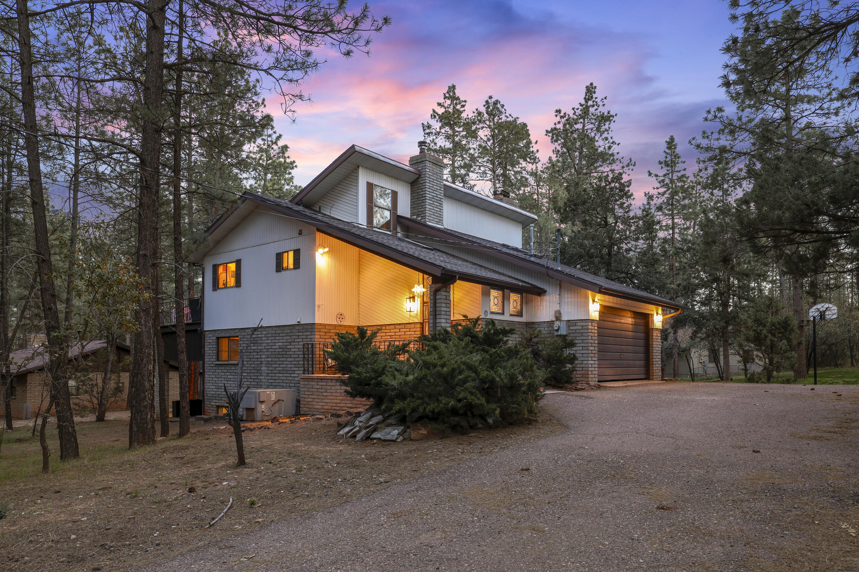 Two-story gray-sided home with an attached garage among tall pines at dusk.