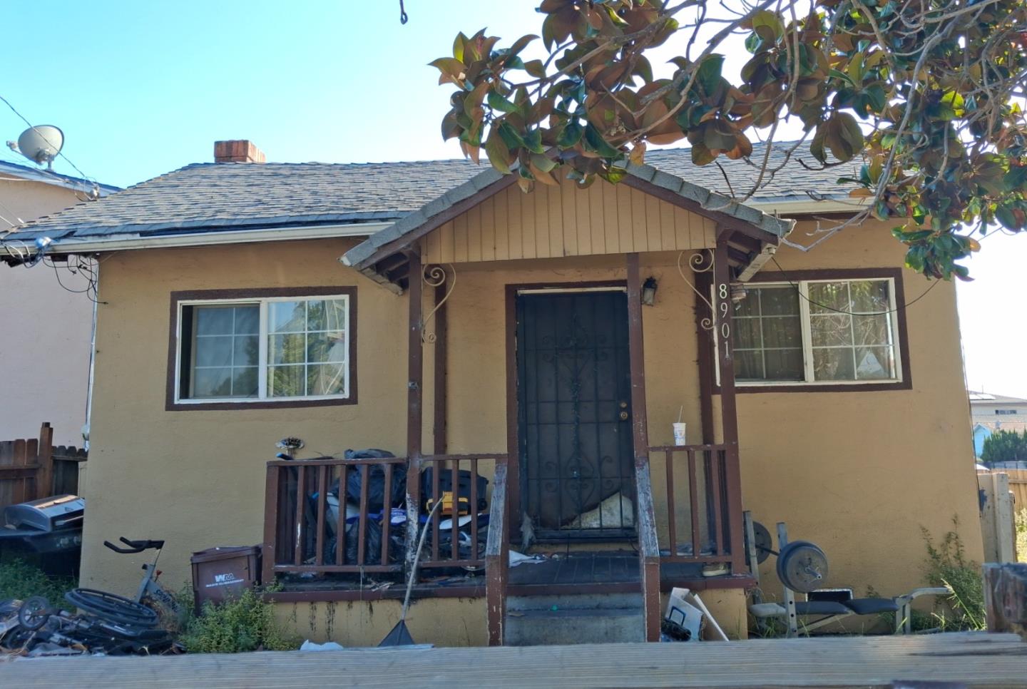 Tan single-story home with a covered porch, dark front door, and cluttered yard.