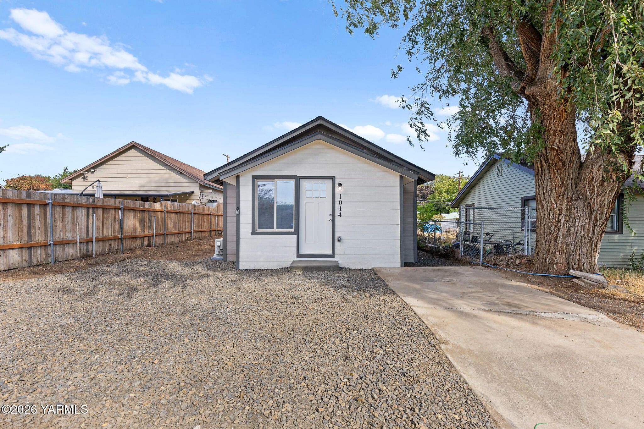 Small light-colored home with dark trim, gravel yard, concrete driveway, and a large mature tree.