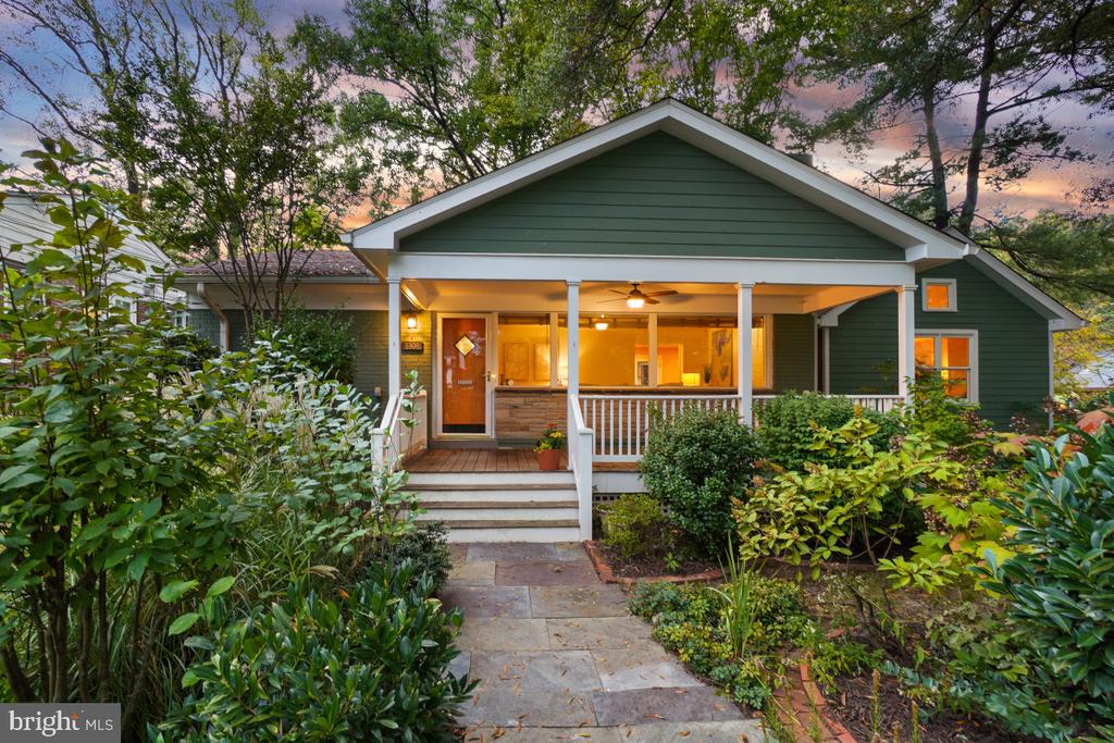 Green house with a covered porch, white railings, and a stone path through lush gardens at dusk.