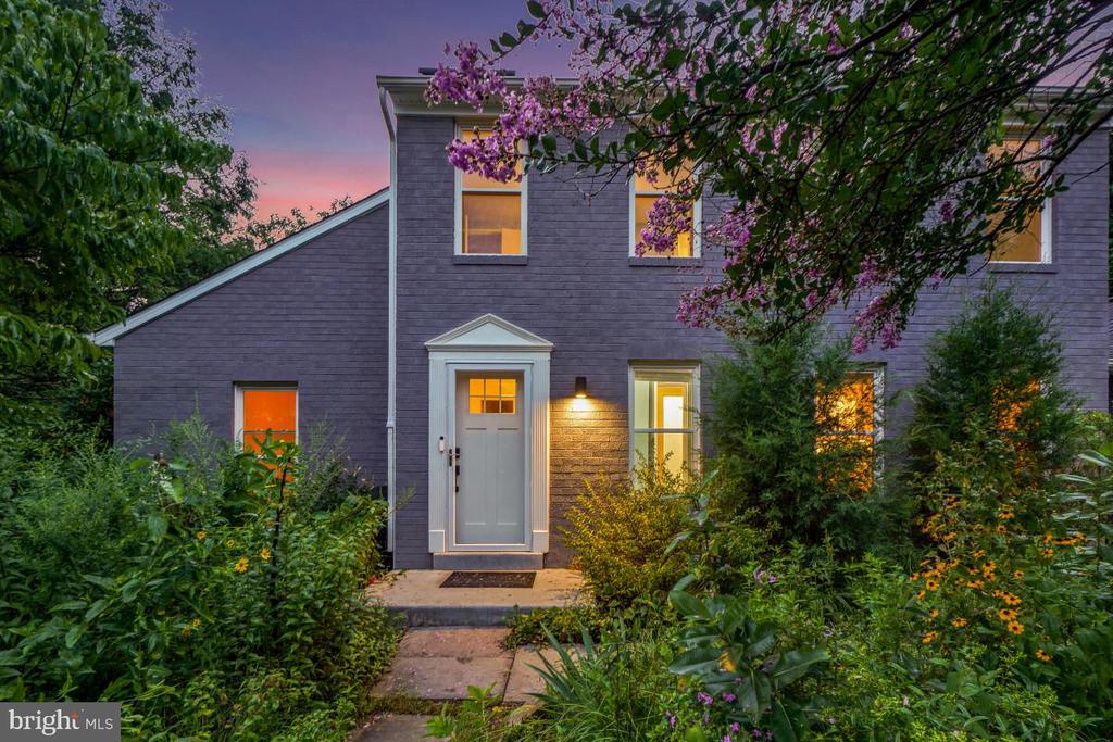 Gray two-story house with a white front door, warmly lit windows, and a lush garden at dusk.