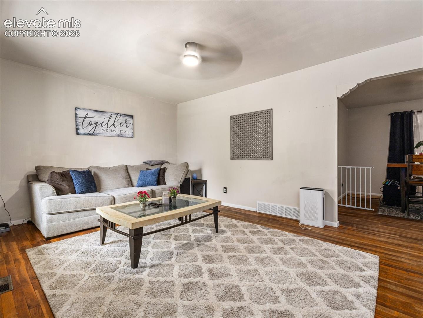 Living room with beige sectional, glass coffee table, patterned rug, and hardwood floors.