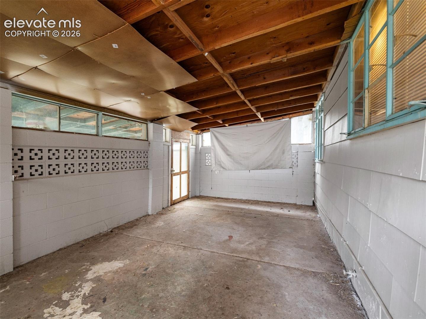 Unfinished enclosed patio with a concrete floor, blue-framed windows, and exposed wooden ceiling rafters.