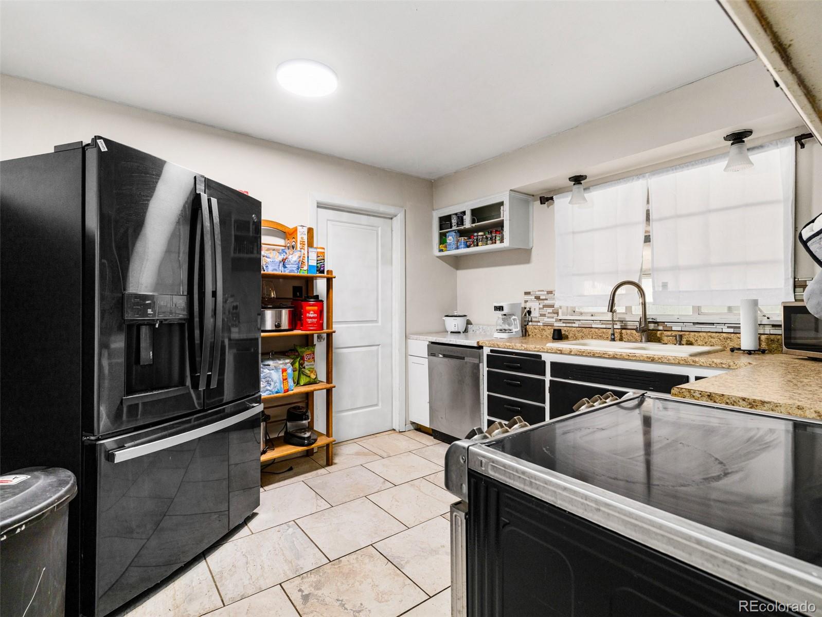 Kitchen with a large black refrigerator, tiled floor, and sink beneath curtained windows.