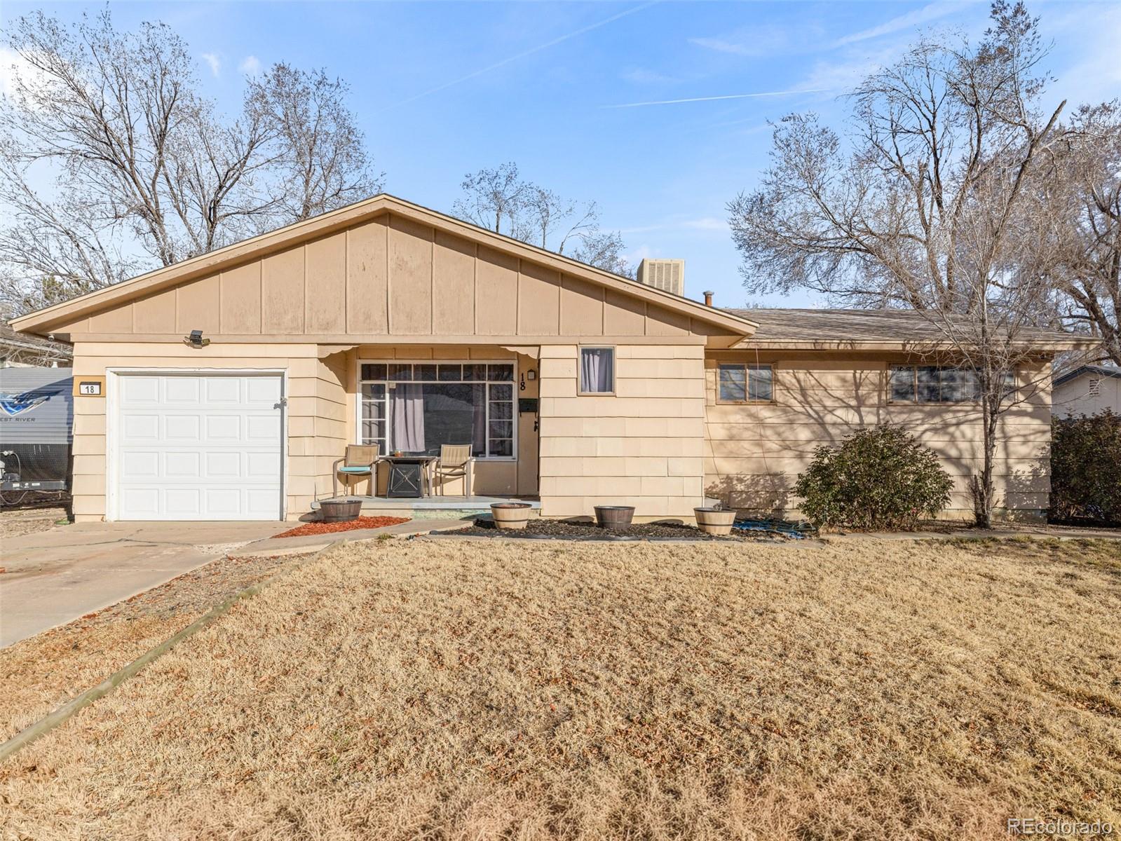Single-story tan home with a white garage door and patio chairs at the covered entry.