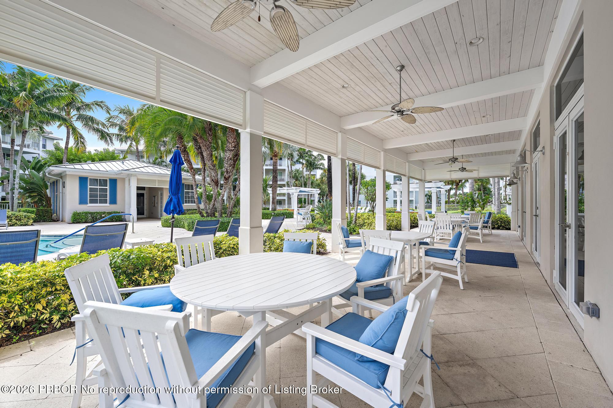 Covered patio with white tables and blue-cushioned chairs beside a pool and palm trees.