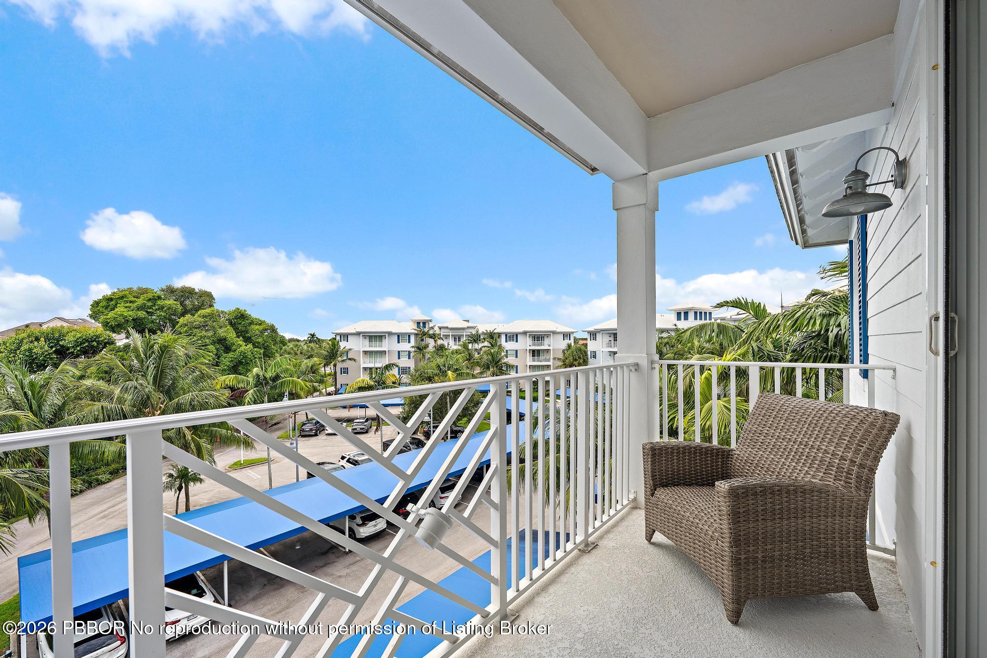 Covered condo balcony with a wicker chair overlooking palm trees and neighboring buildings.