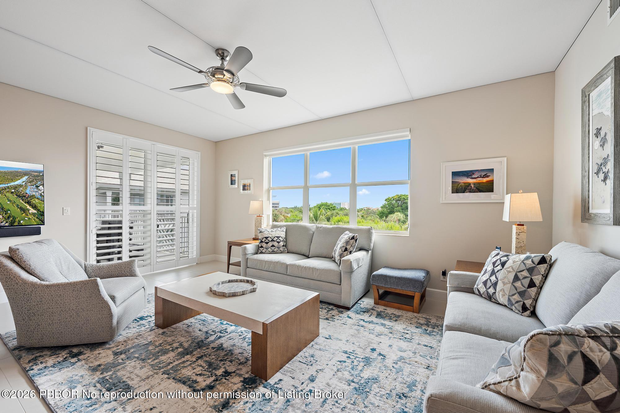 Furnished living room with gray sofas, a patterned rug, plantation shutters, and a large window overlooking greenery.