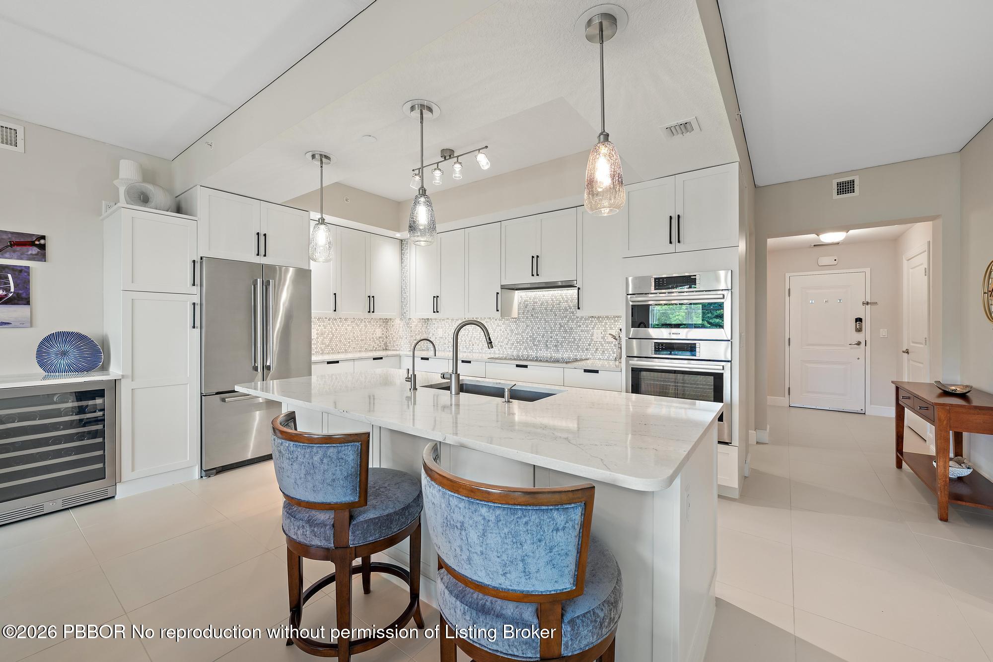 Modern kitchen with a large white island, blue stools, stainless appliances, and pendant lights.