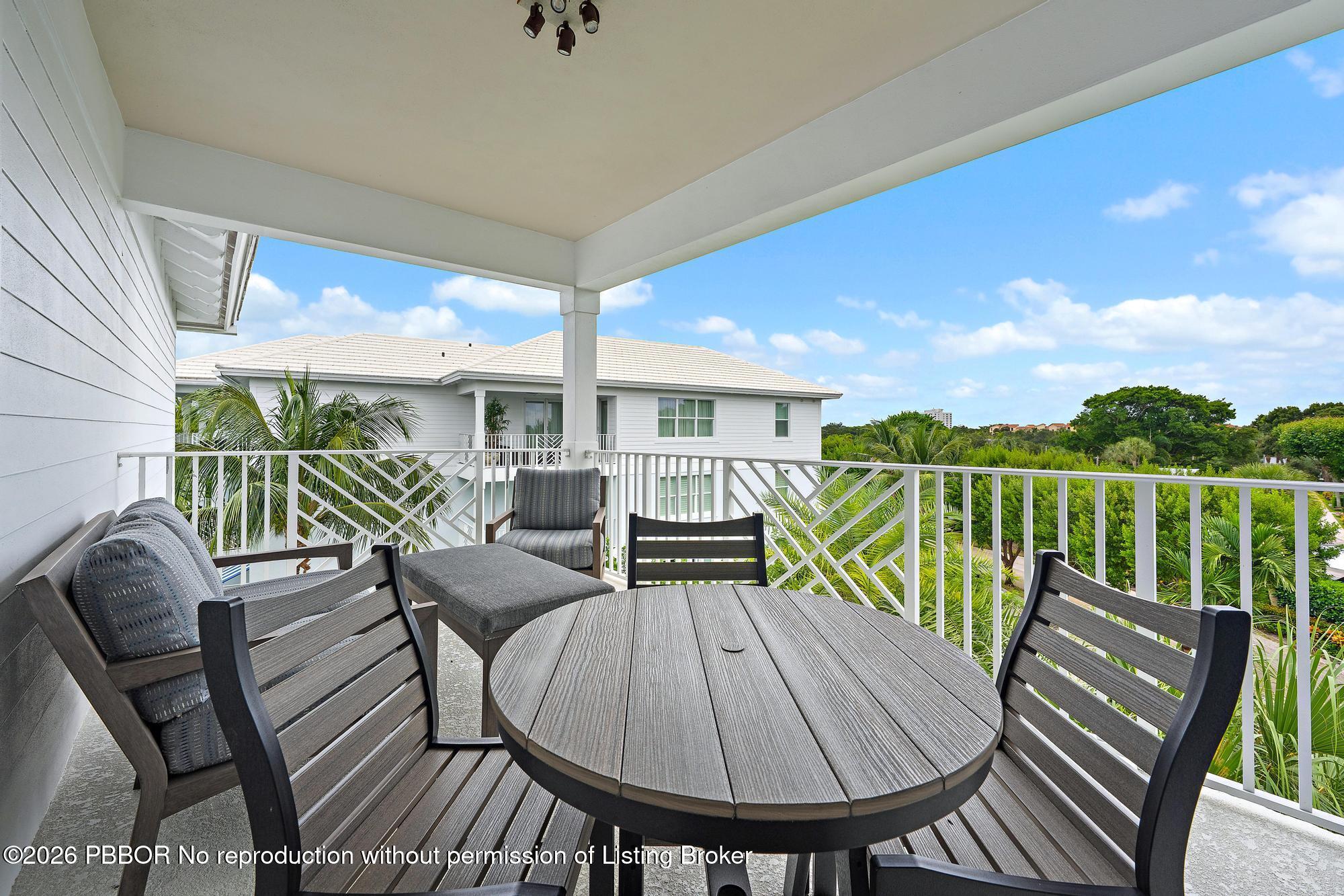 Covered balcony with a round dining table, dark chairs, and views of palms and greenery.