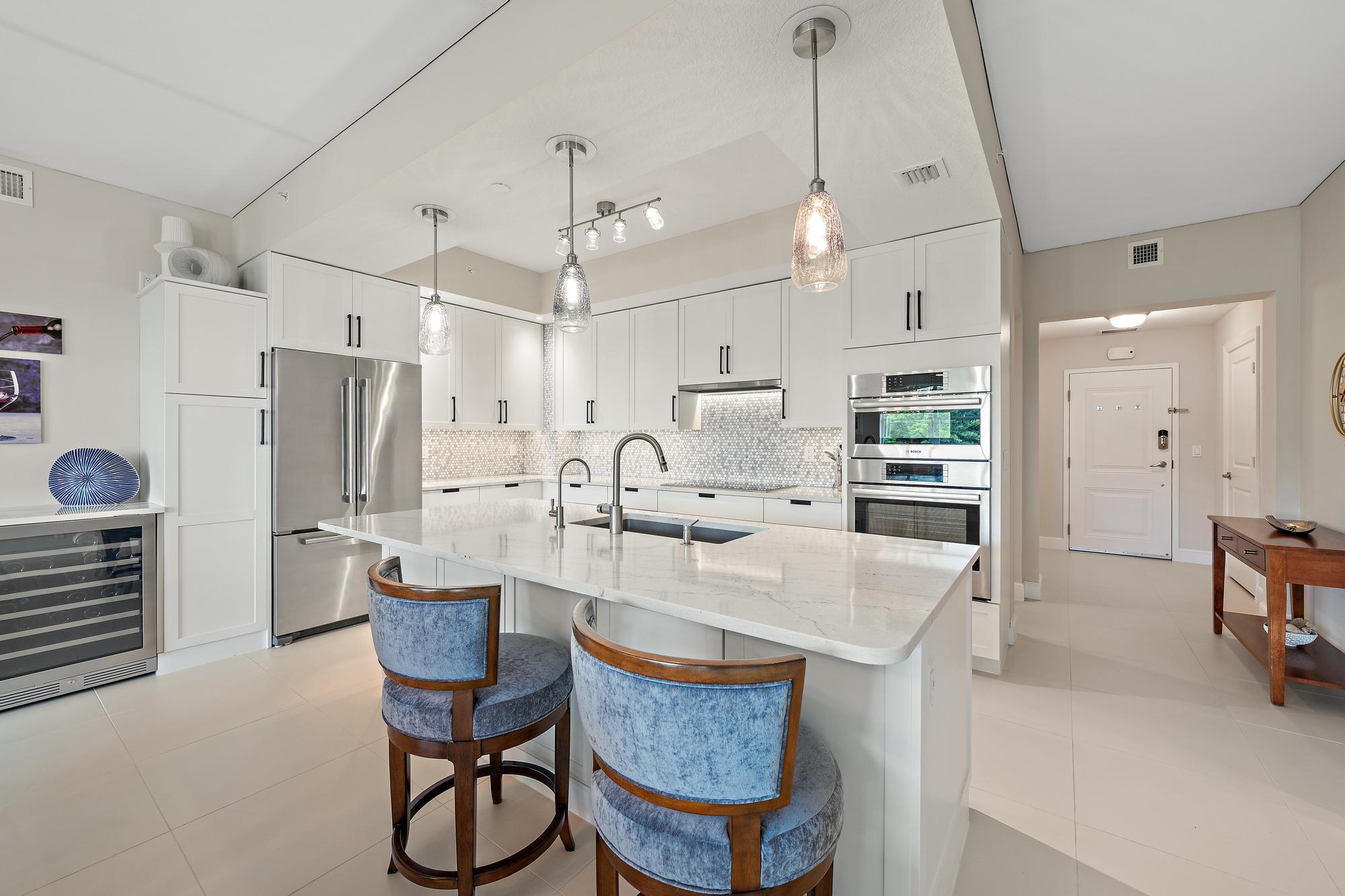 Modern white kitchen with a large island, blue bar stools, stainless appliances and pendant lights.