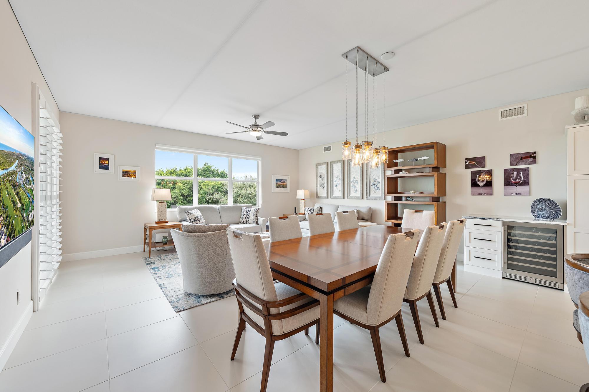 Dining and living area with a large wooden table, upholstered chairs, and windows overlooking greenery.