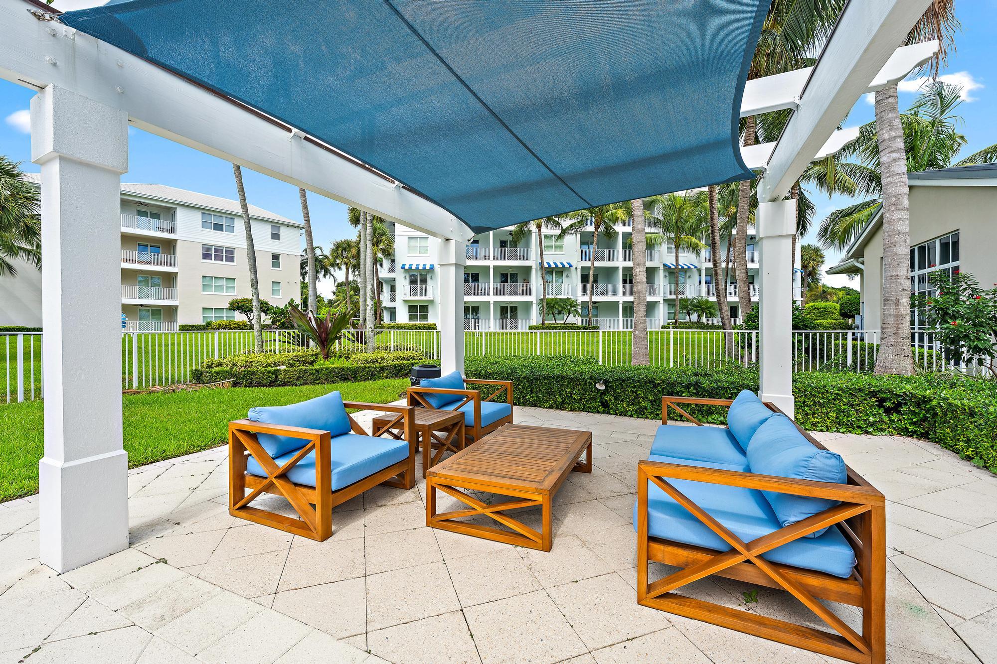 Patio with blue-cushioned wooden seating beneath a shade canopy, overlooking a lawn and palm trees.
