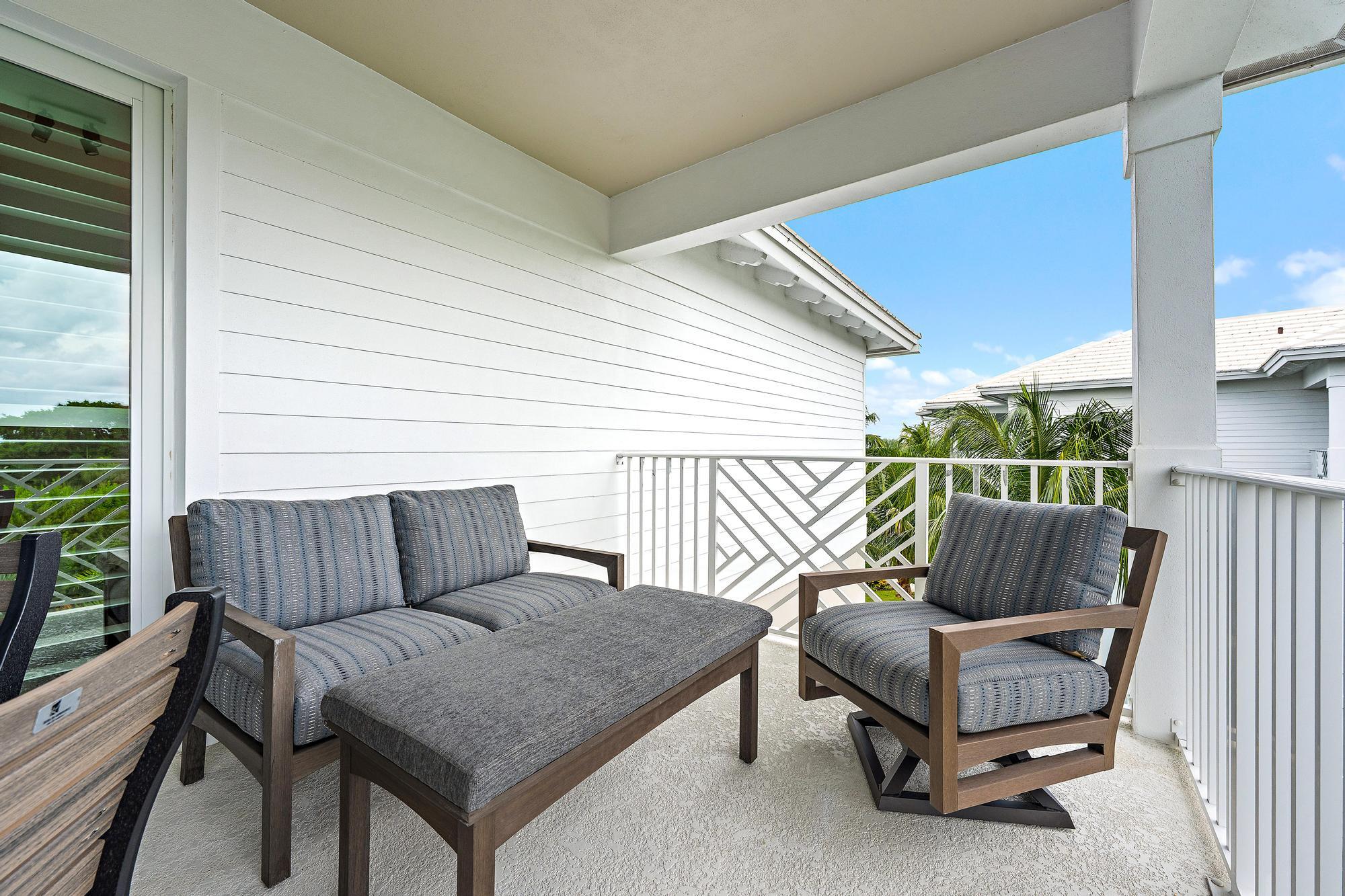 Covered balcony furnished with gray outdoor seating and a coffee table, overlooking palm trees.