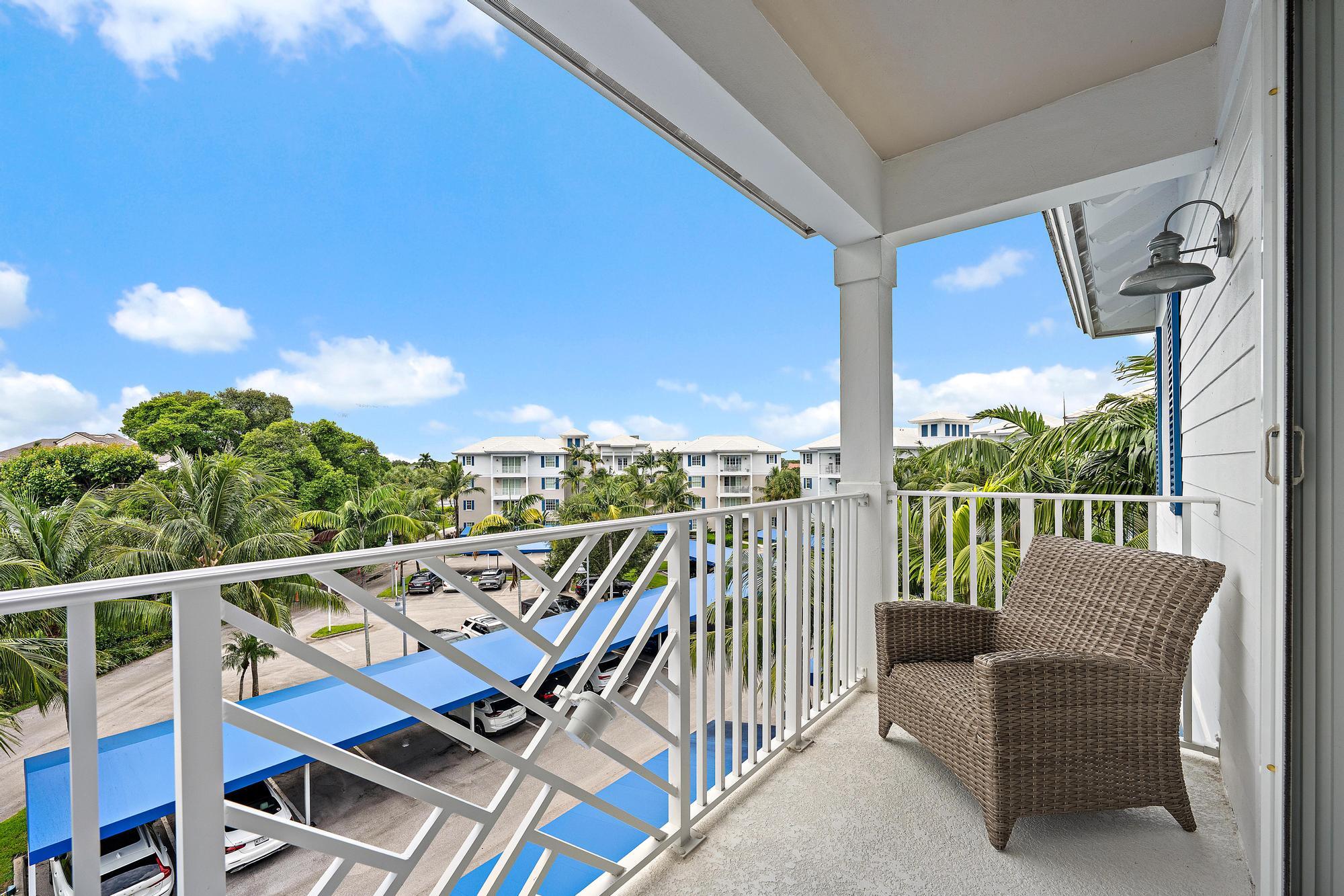 Covered balcony with a wicker chair overlooking palm trees and neighboring buildings.