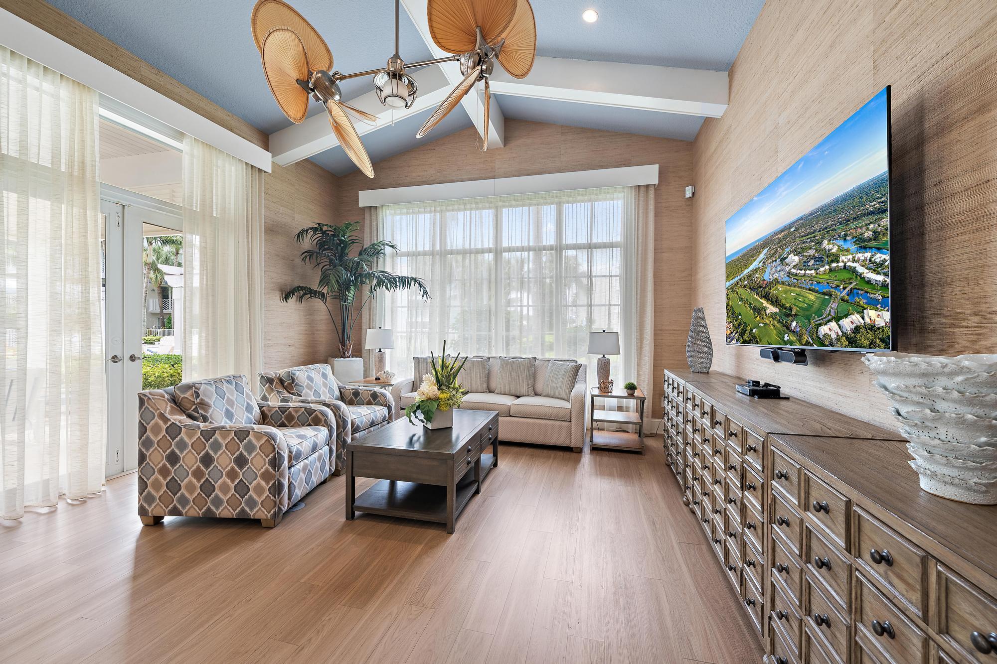 Living room with a vaulted ceiling, patterned armchairs, sheer-curtained windows, and a television above a long wood dresser.