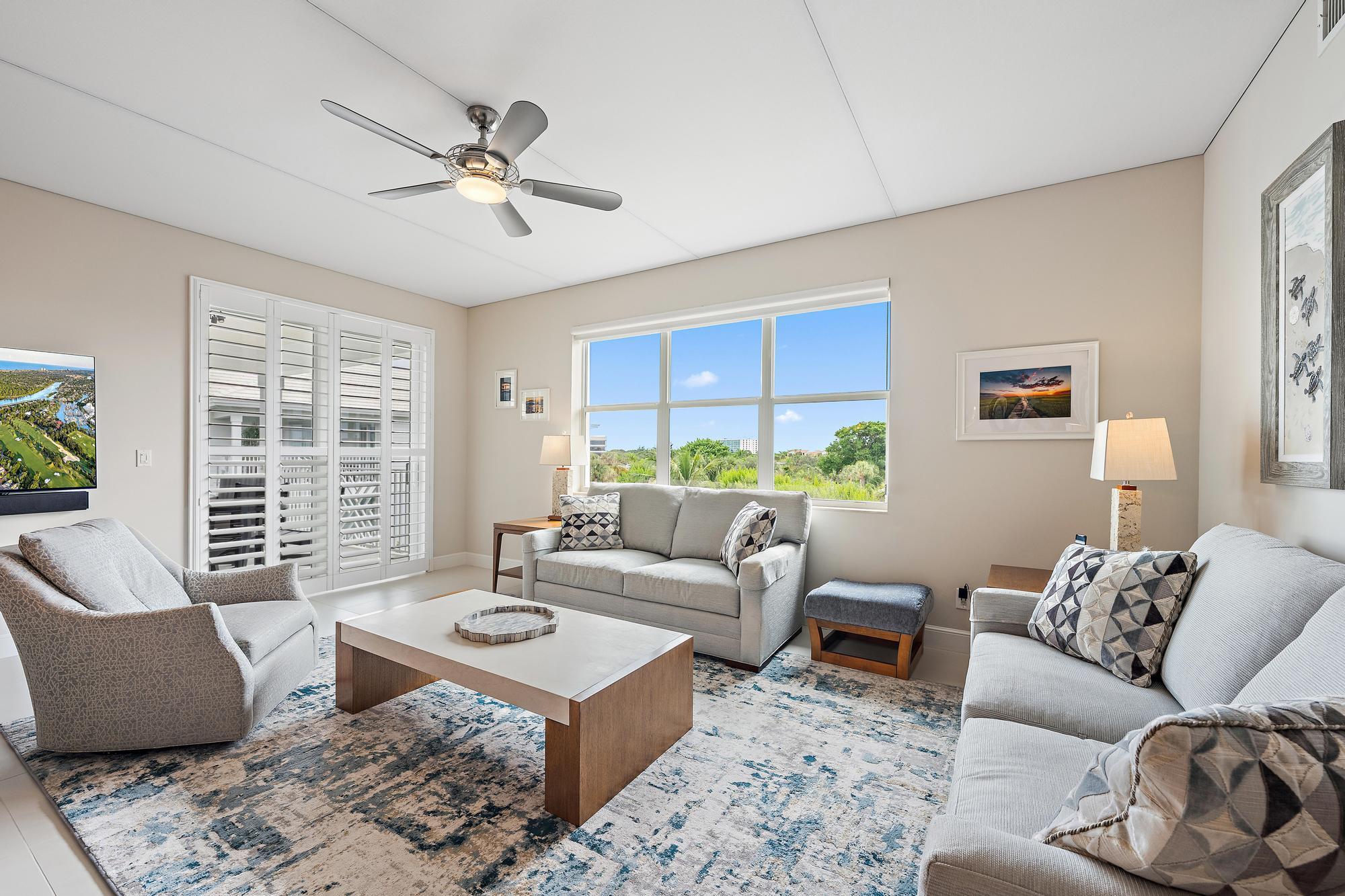 Furnished living room with plantation shutters, a large window, and pale seating around a coffee table.